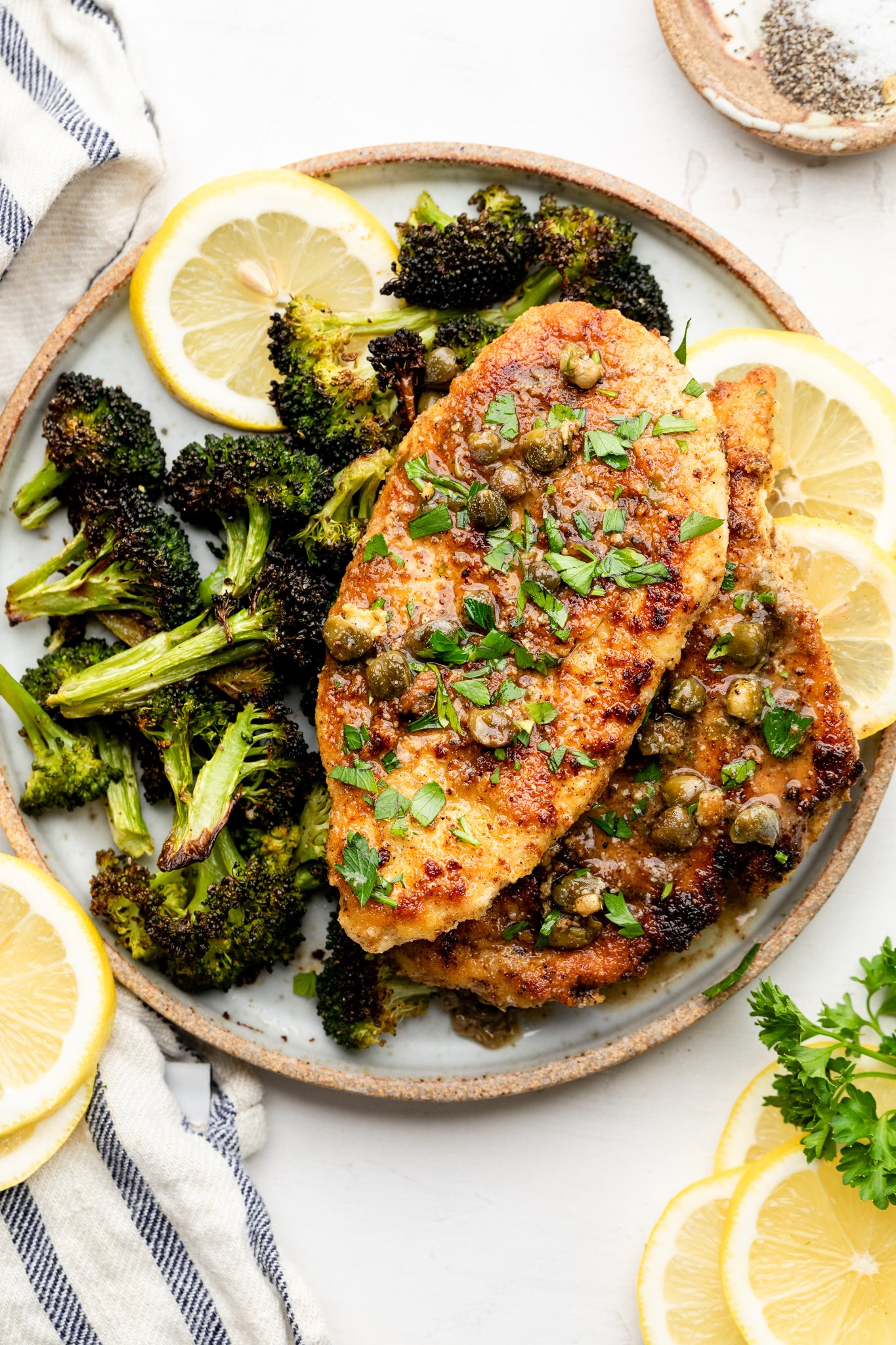 A tan and white plate with roasted broccoli, gluten free chicken piccata, and lemon slices