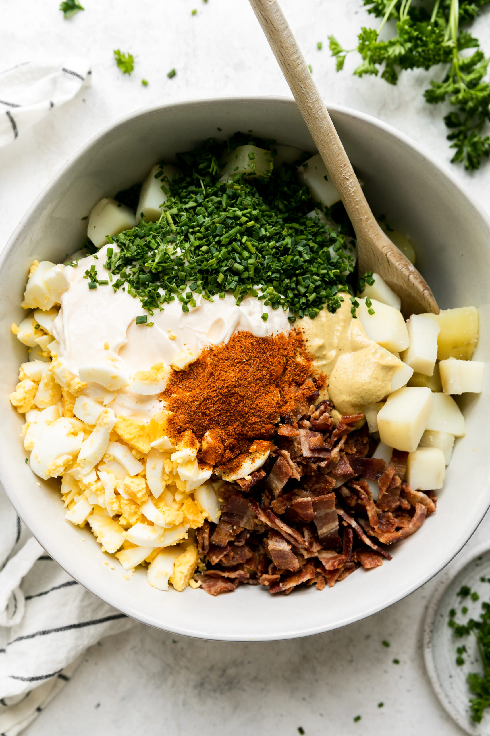 a white bowl with ingredients that have not been mixed: cubed potatoes, chopped hard boiled eggs, chives, parsley, spices, and bacon with a wooden spoon