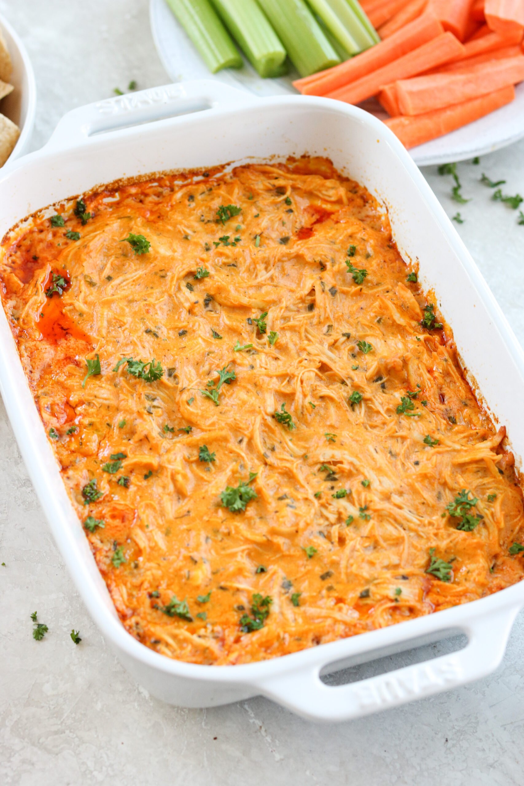 Healthy buffalo chicken dip in a white Staub baking dish topped with parsley with carrots and celery in the background