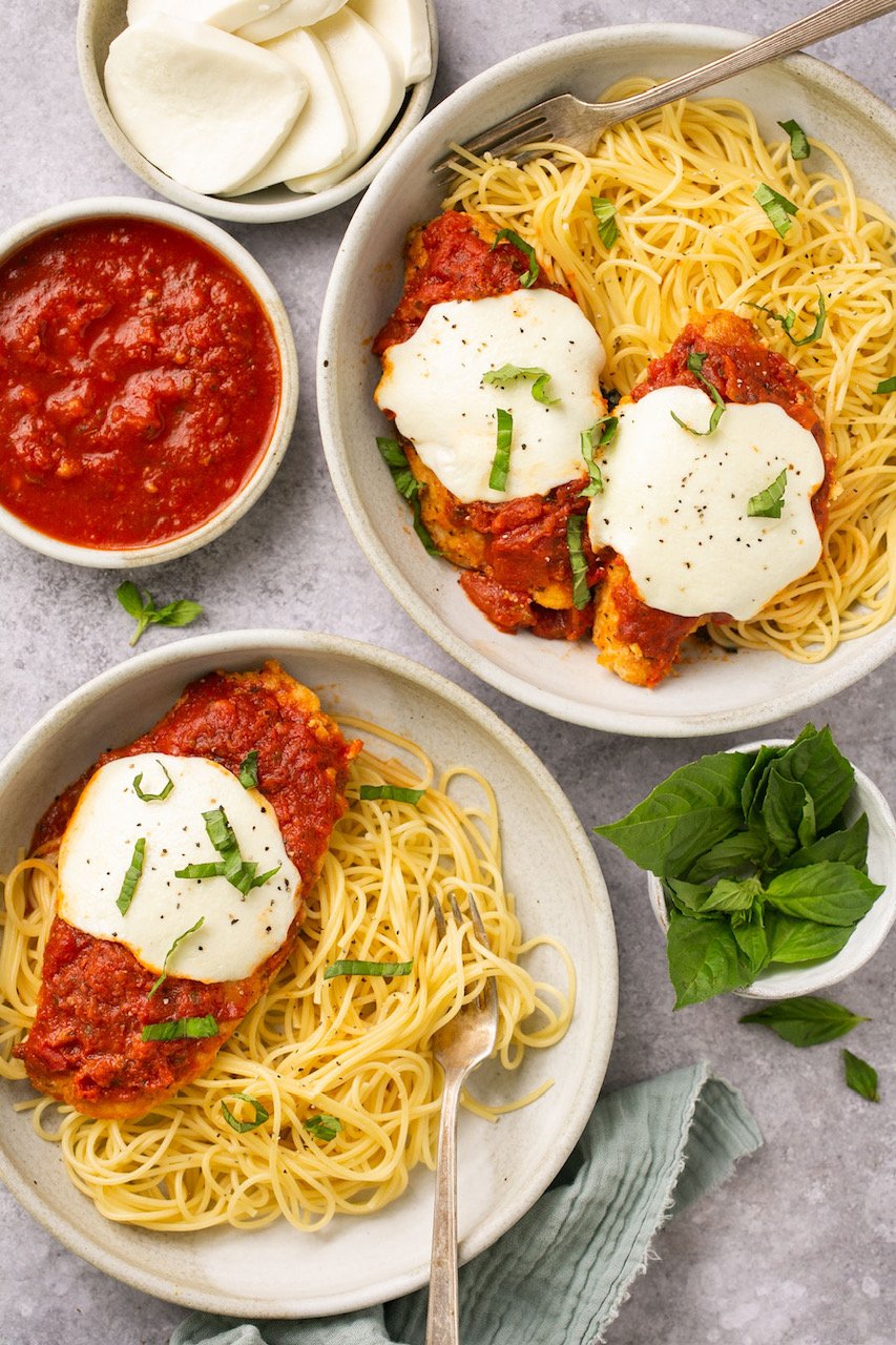 2 bowls of pasta topped with chicken parmesan surrounded by sauce and cheese in nesting bowls