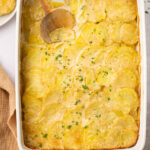gluten free scalloped potatoes in a casserole dish with a wooden spoon taking a scoop out