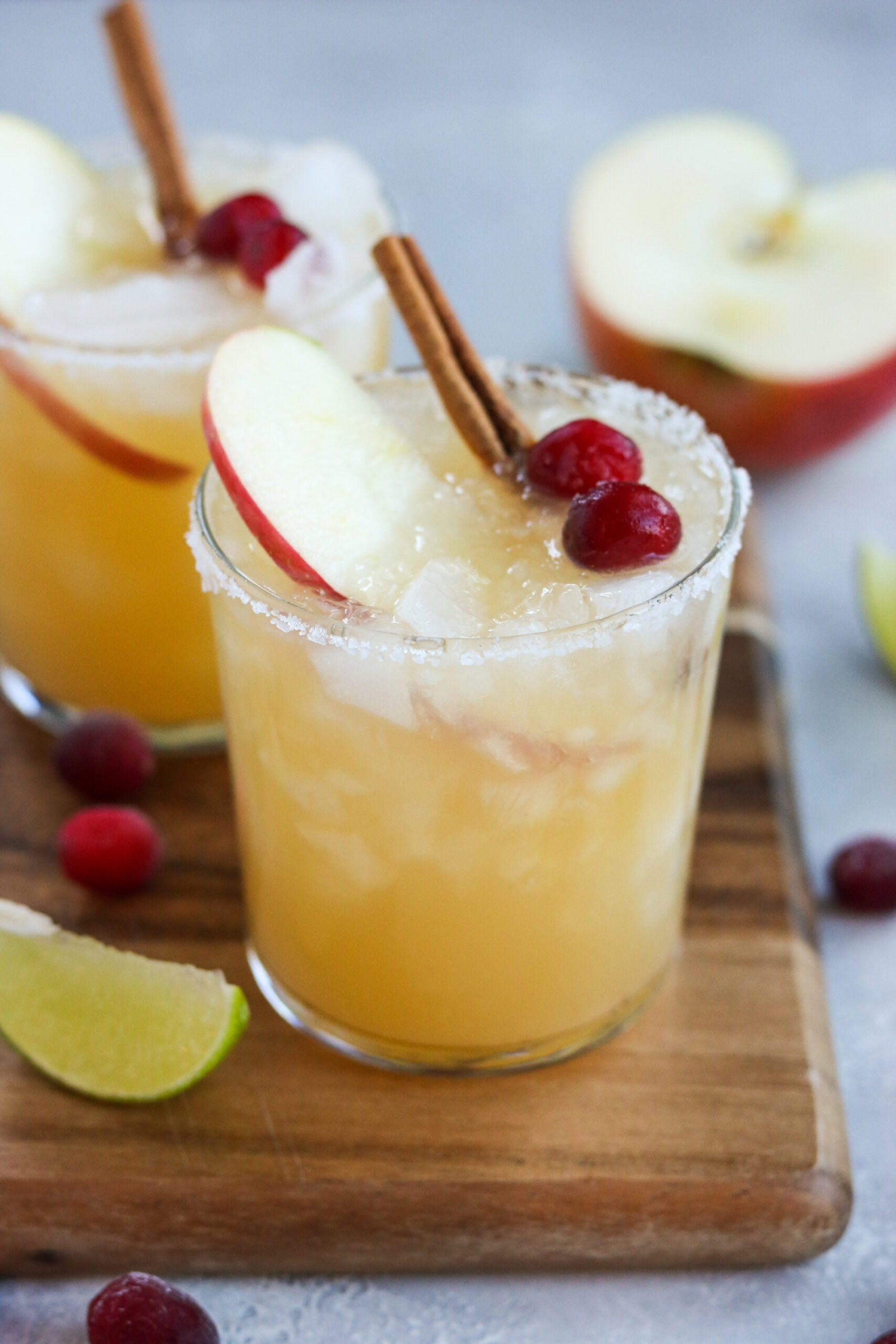 a highball glass filled with ice and an apple cider margarita mixture. Topped with an apple slice, two cranberries and a cinnamon stick with a sea-salt rim
