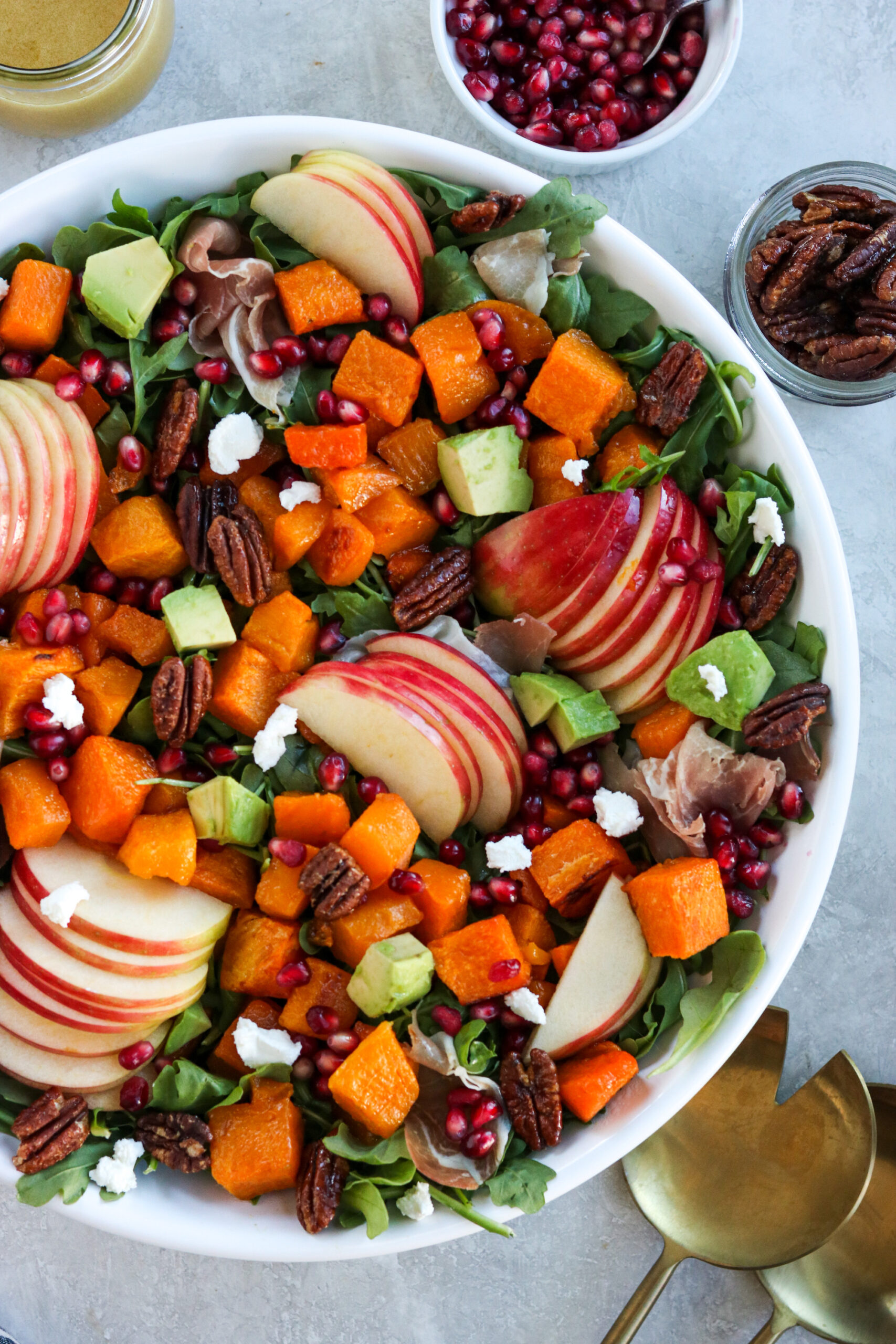 Autumn Harvest Salad win a white bowl surrounded by spoons, pomegranate seeds, pecans, and dressing