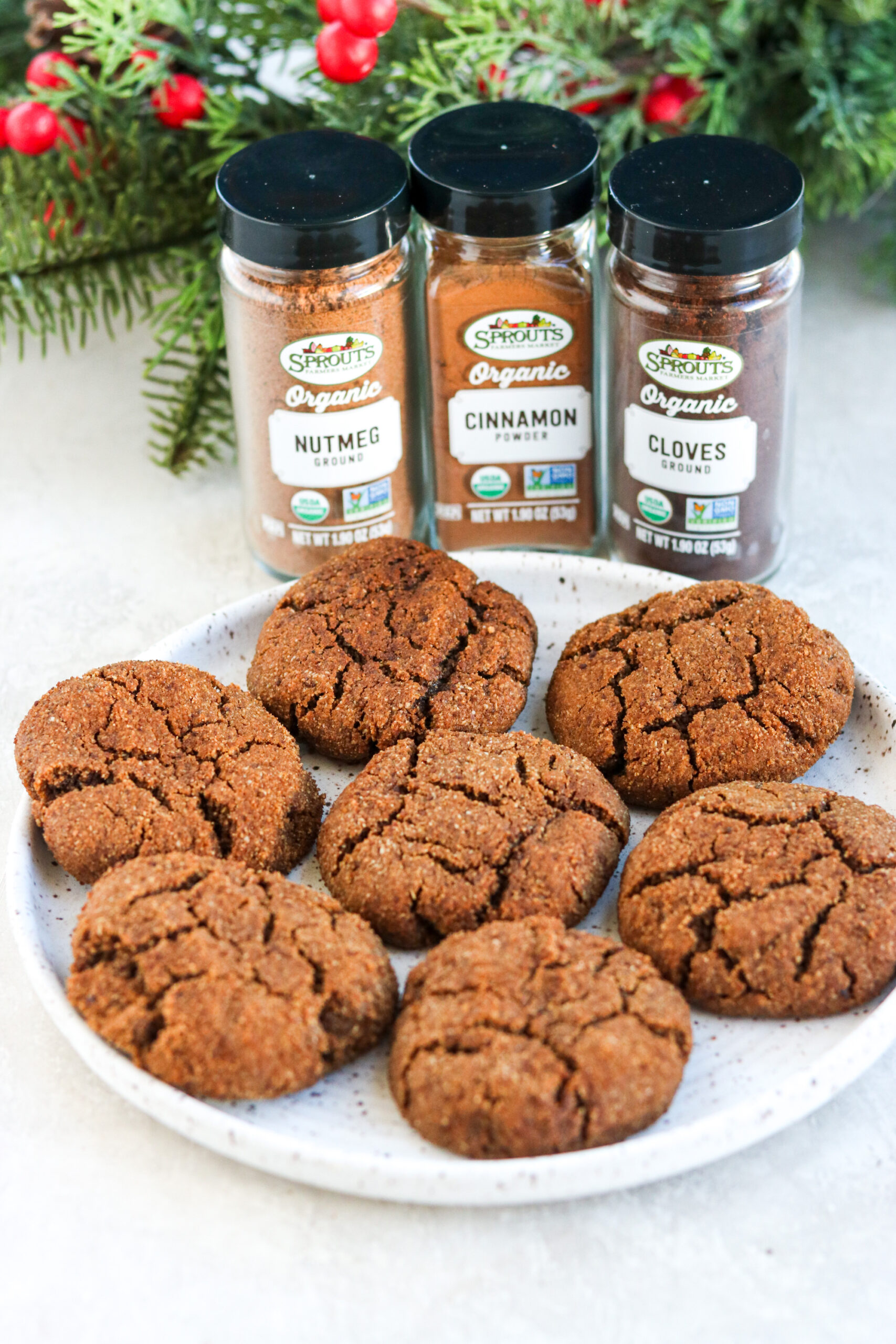 seven baked ginger cookies on a white plate with nutmeg, cinnamon, and cloves in the background