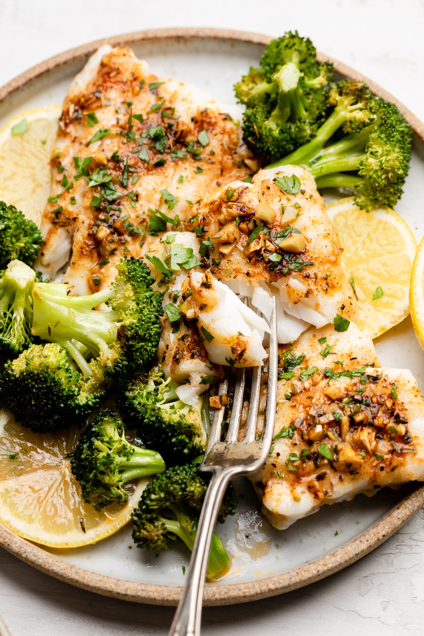 cajun cod with broccoli garnished with parsley and sliced lemons on a white plate with a silver fork 