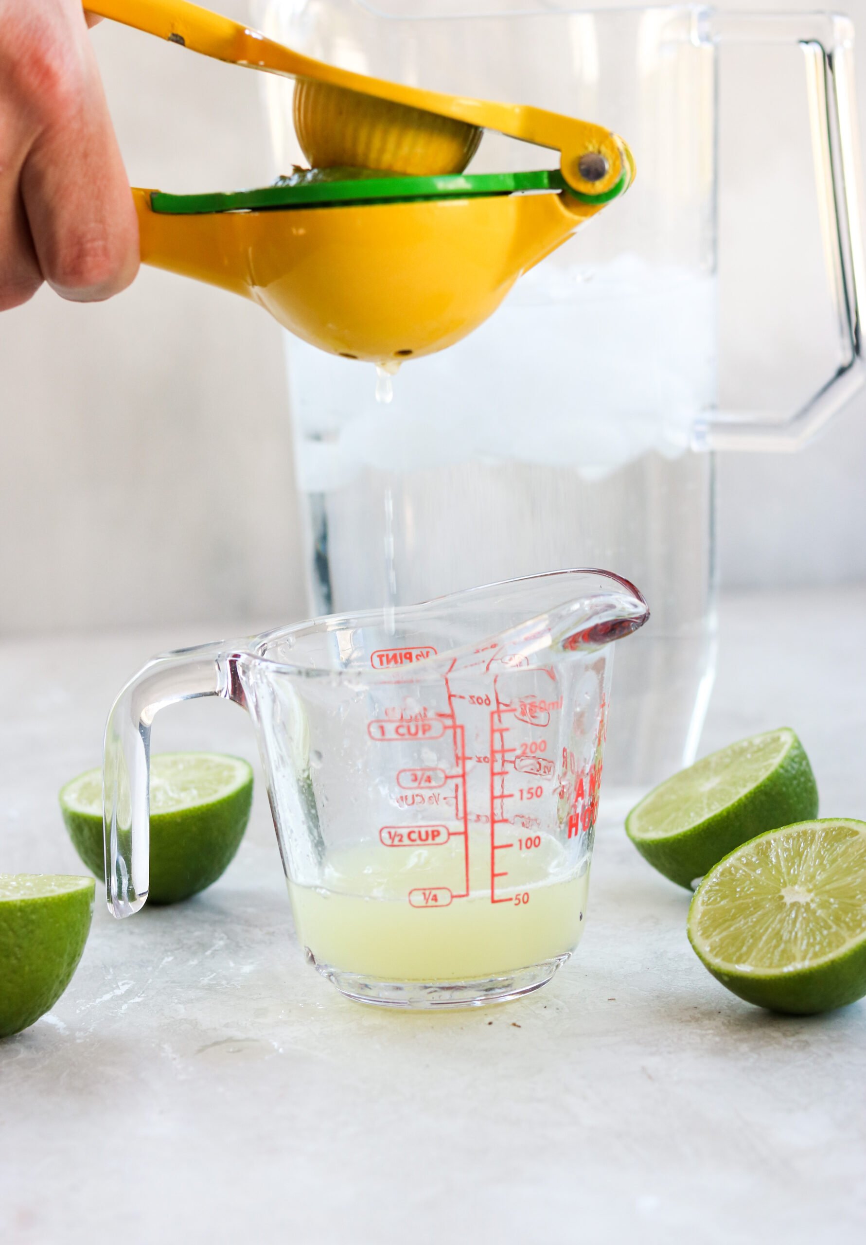 a hand using a metal citrus juicer to squeeze lime juice into a pyrex measuring cup next to three lime halves. 