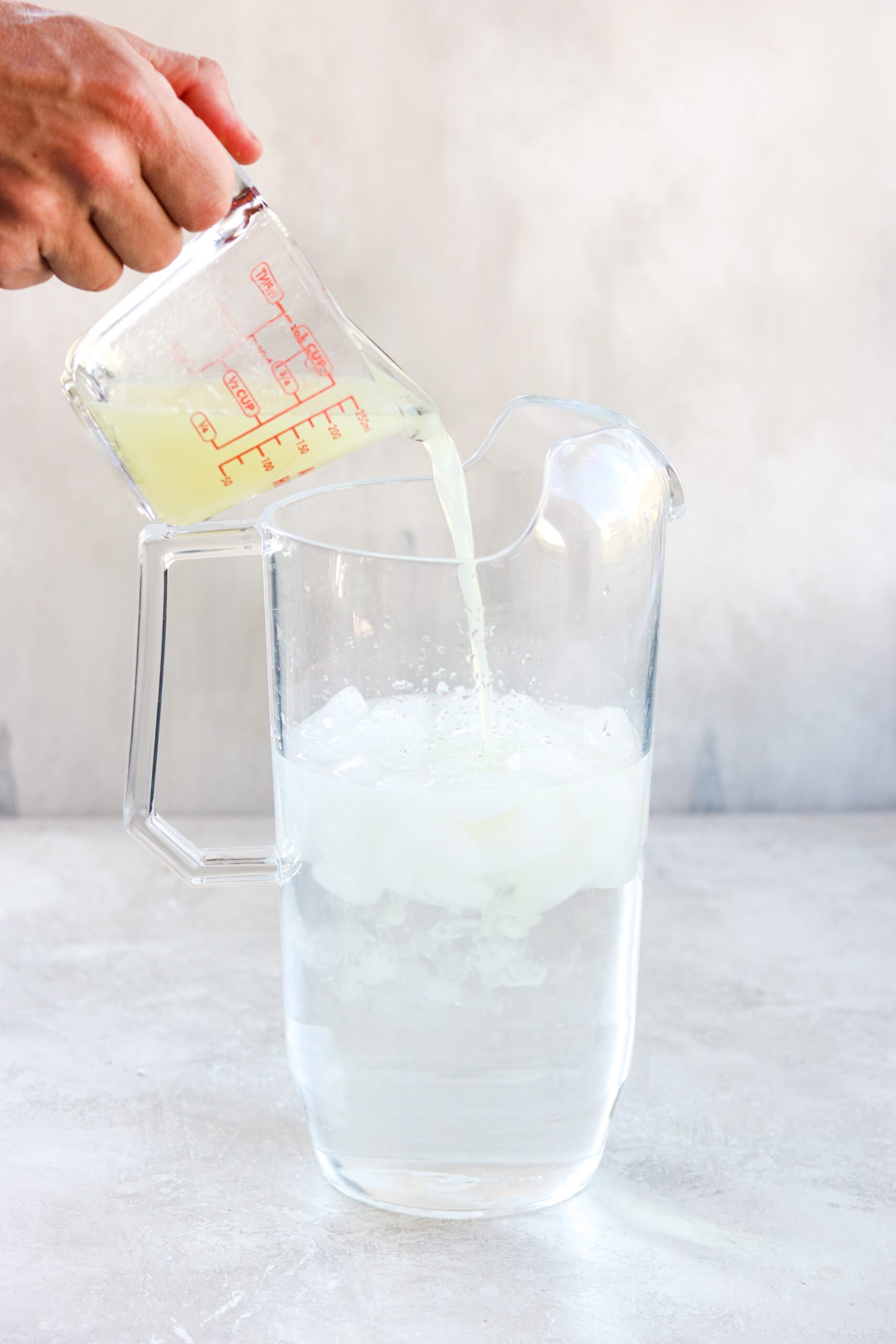 a pyrex measuring pitcher pouring lime juice into a clear pitcher filled with ice and cold water. 