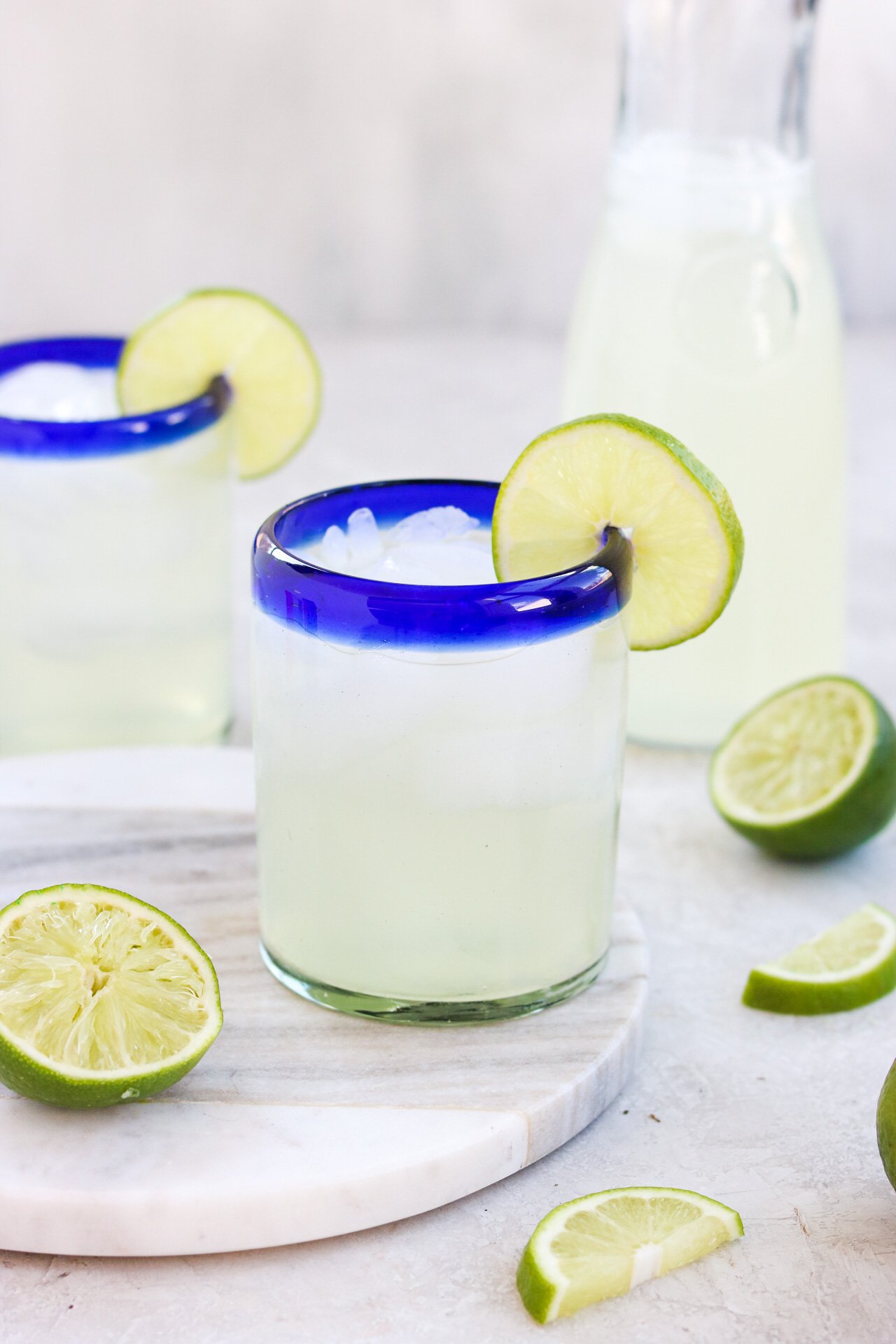 a closeup view of one glass filled with ice and limeade with a lime wedge on the rim with a squeezed lime next to the glass. Another glass of the same is in the background.