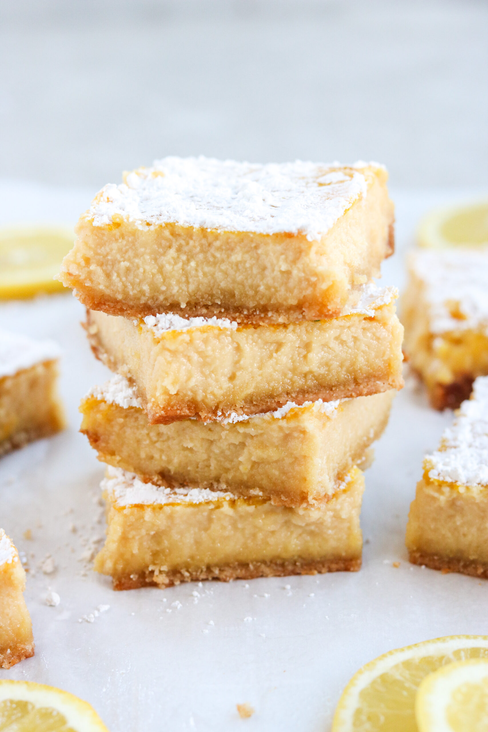 a close up of a four lemon bars stacked on top of each other with lemon slices on white parchment paper
