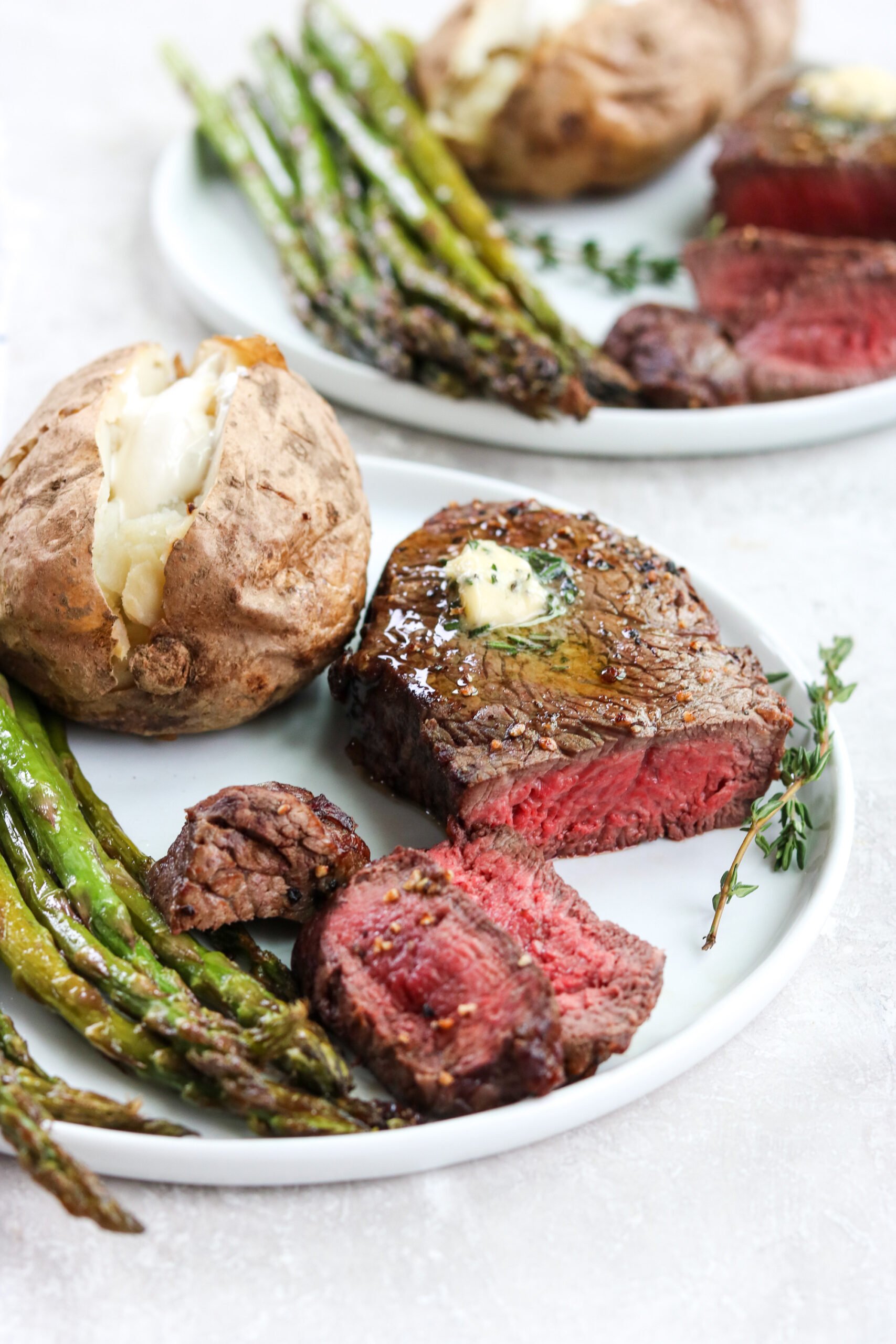 two white plates with a baked potato, sliced air fryer filet mignon, and asparagus.