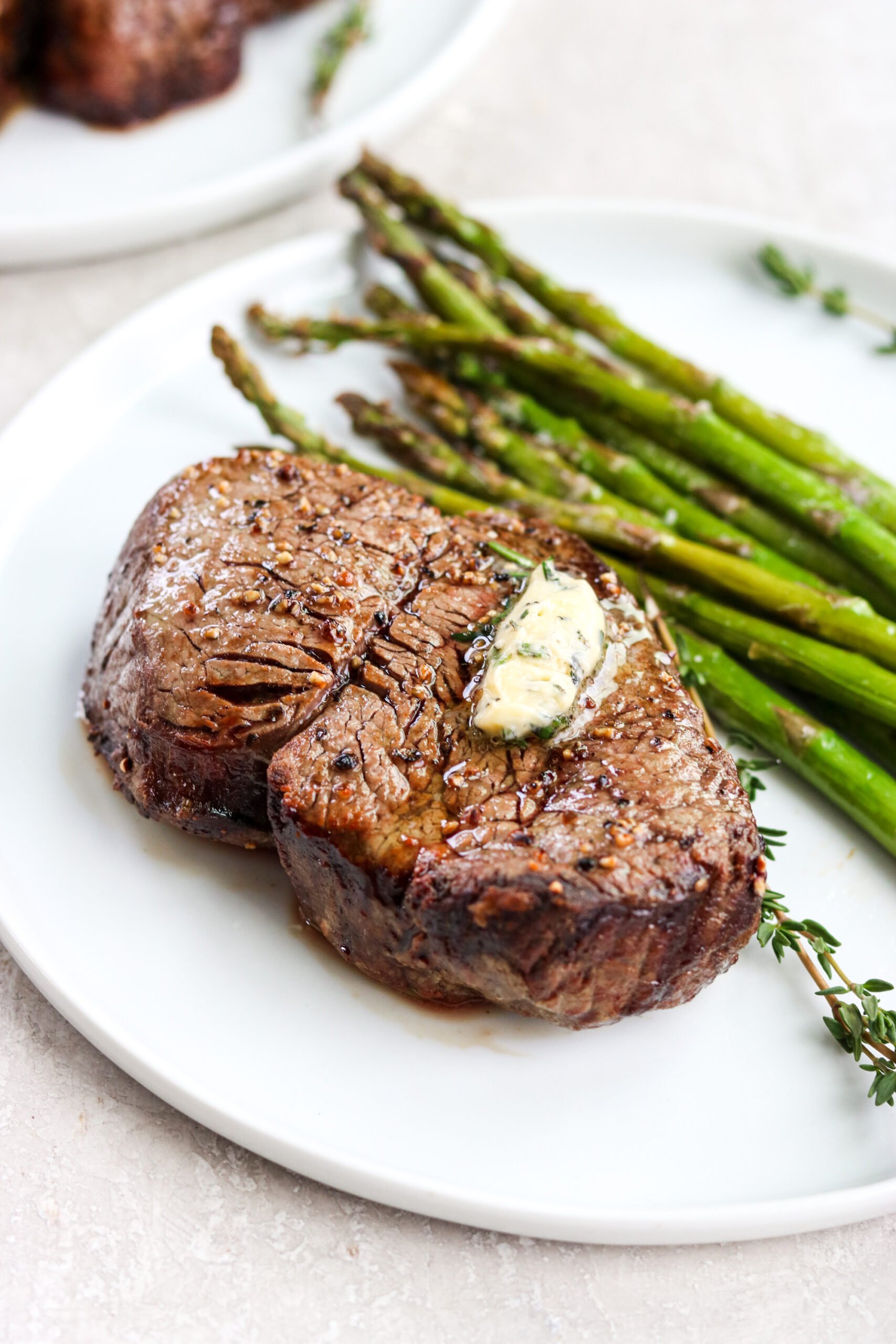 a cooked steak with herb butter on top next to grilled asparagus on a white plate. 