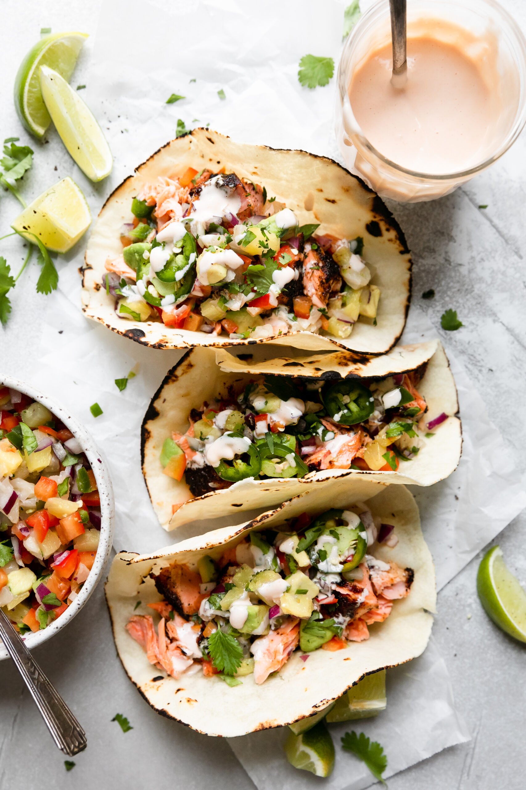 three small tortillas filled with salmon, pineapple, salsa, and lime aioli, next to a glass jar of aioli, a bowl of salsa, and three lime wedges. 