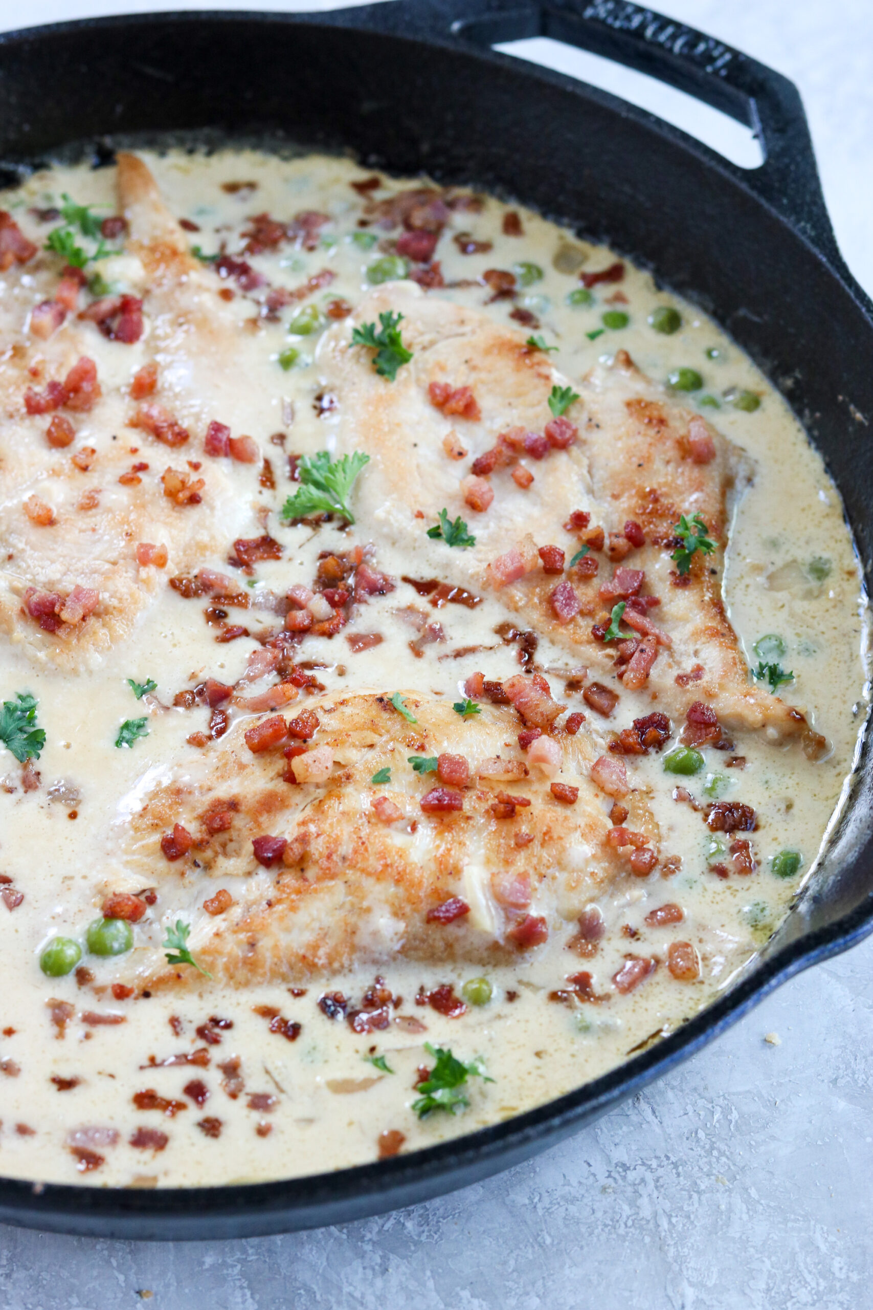 a closeup photo of a cast iron pan with cooked chicken breasts, peas, creamy sauce, and topped with crispy pancetta and parsley 