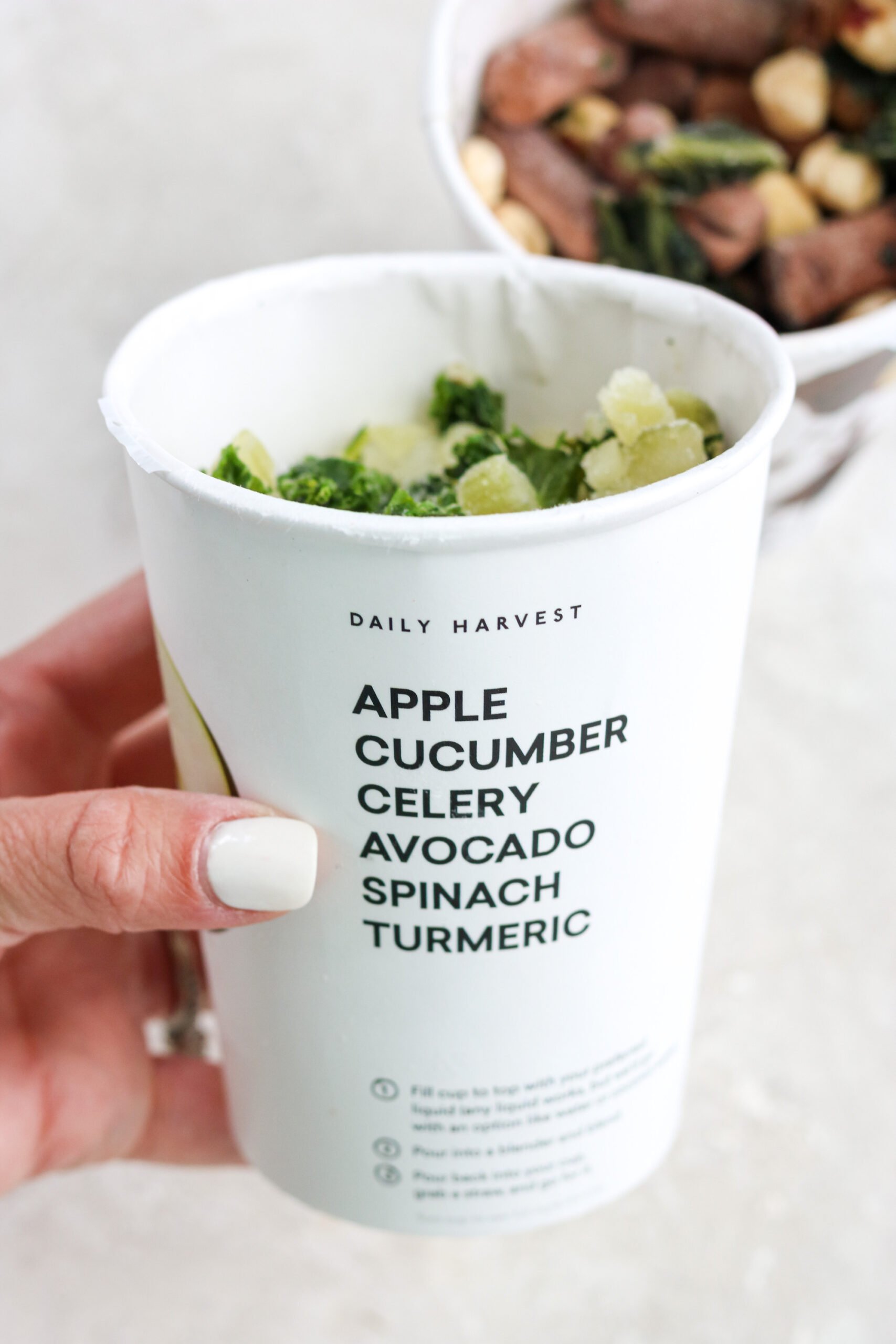 A Daily Harvest Apple veggie smoothie before blending in a white cup with the ingredients written on the side of the cup in black