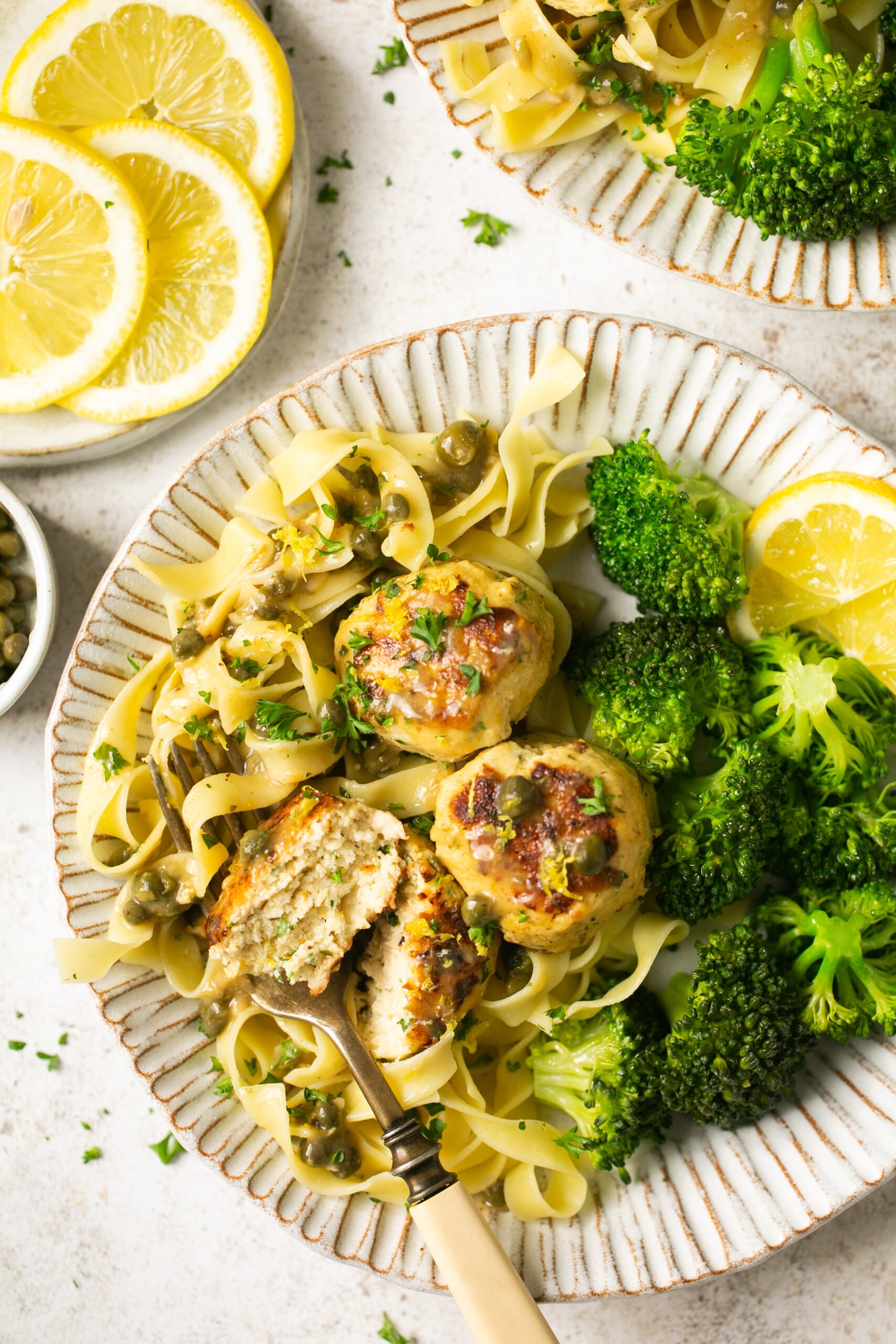 chicken piccata meatballs with gluten free pasta and broccoli on a white scalloped plate with parsley and lemon