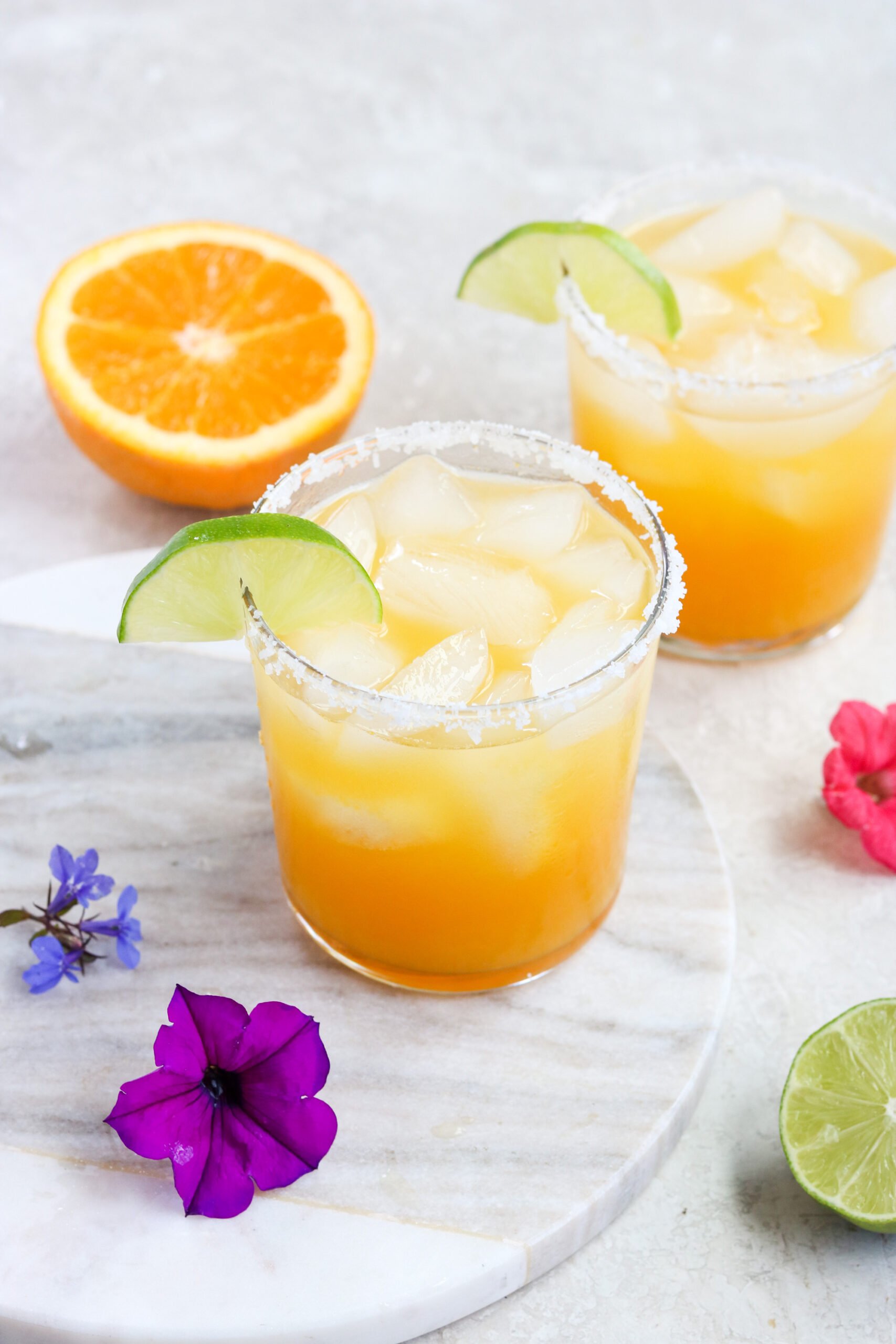 two clear glasses filled with passion fruit margaritas next to half an orange and purple flowers