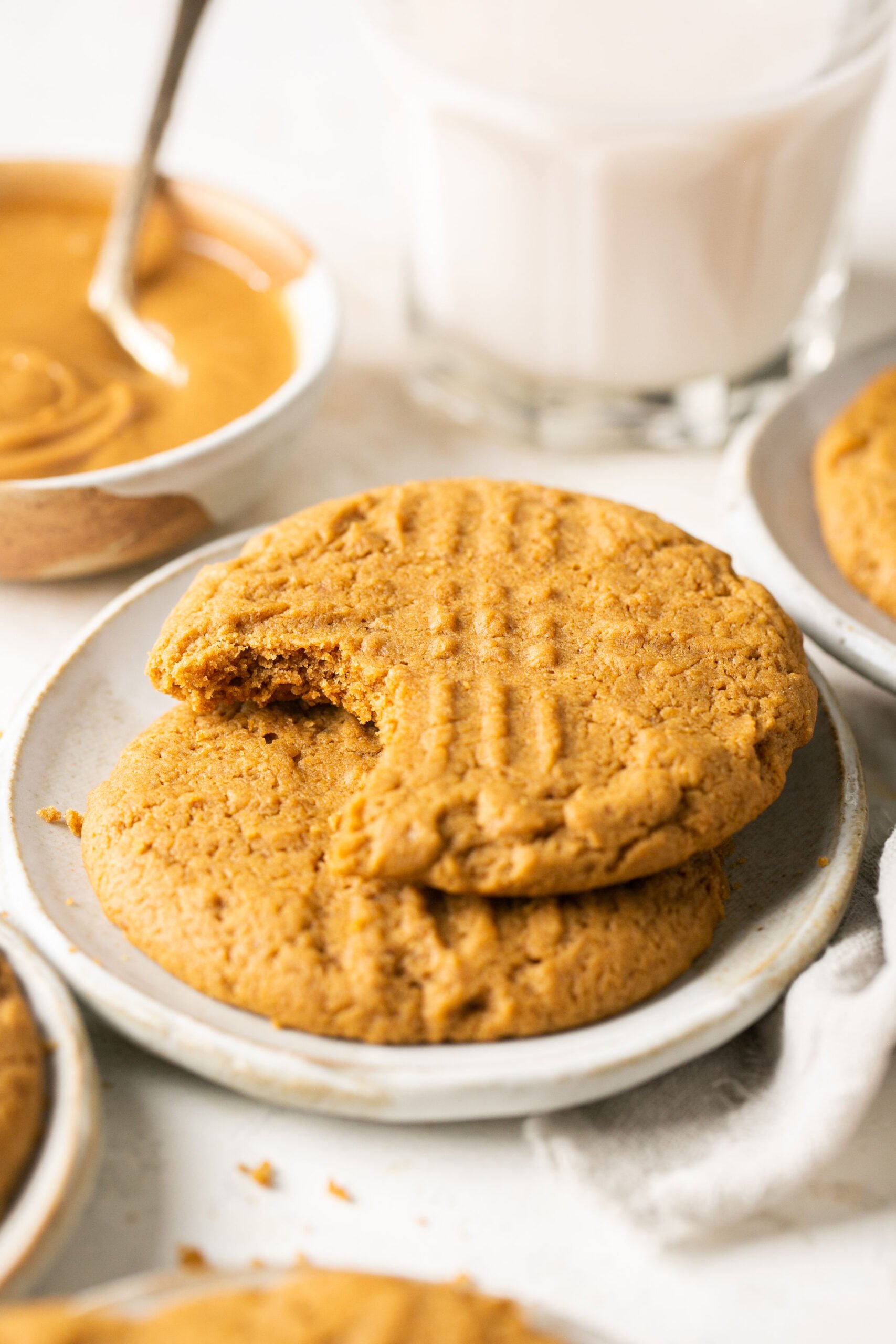 2 peanut butter cookies stacked on a white plate with a bowl of creamy peanut butter in the background