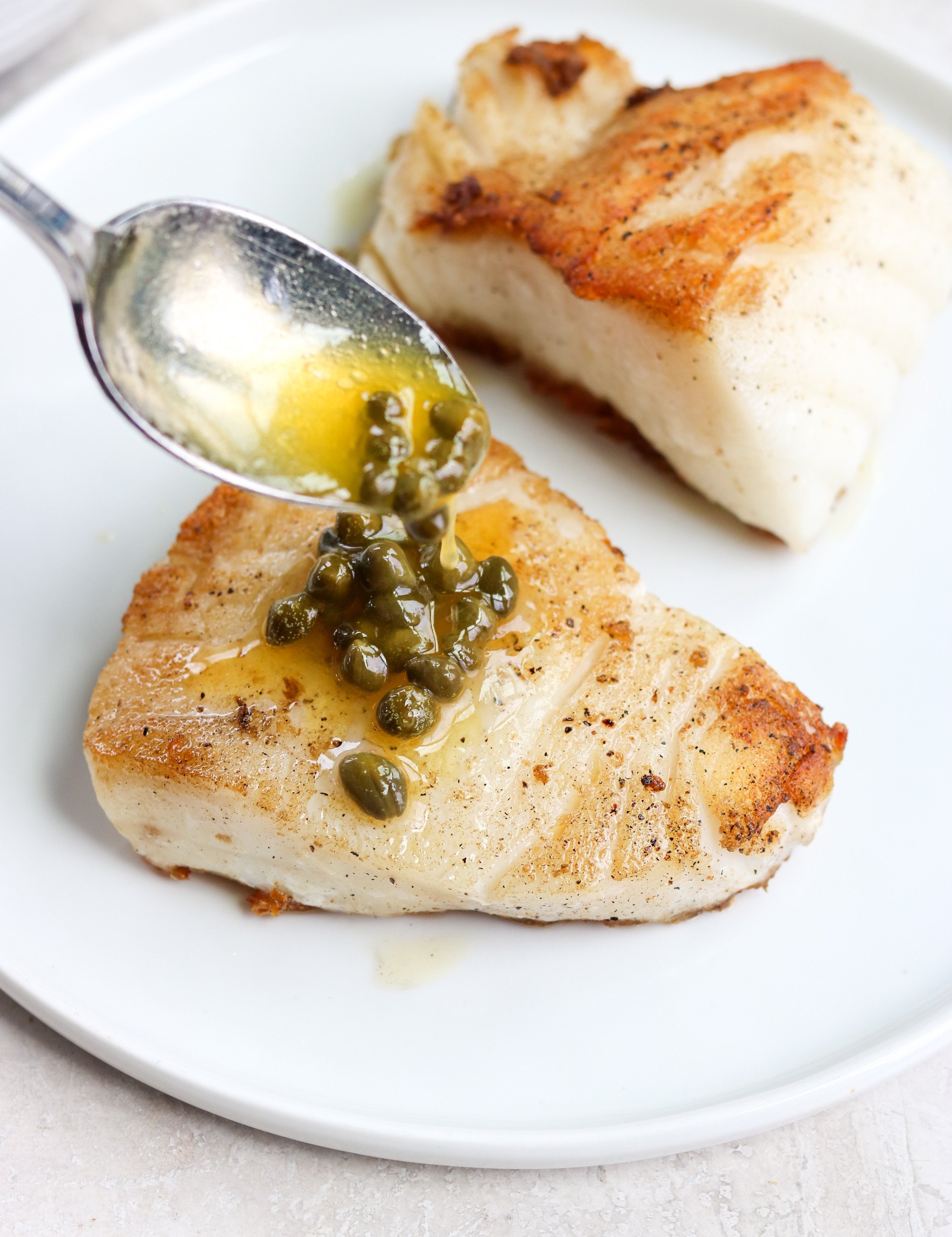 a silver spoon drizzling lemon caper butter over a piece of Chilean sea bass on a white plate