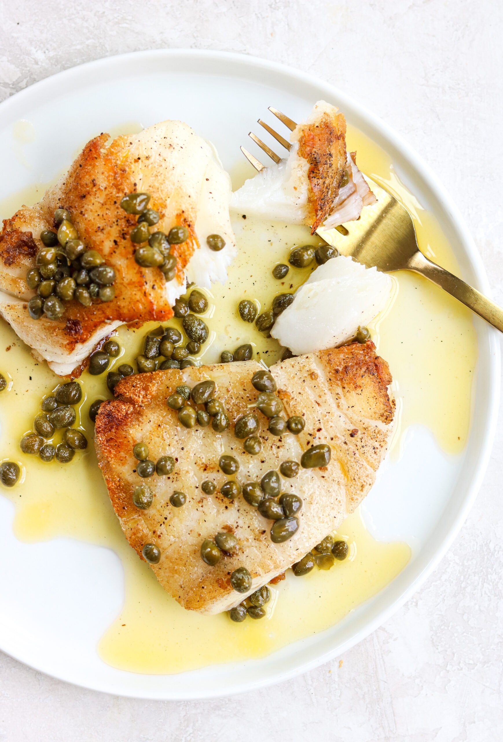 crispy pan seared white fish on a white plate with lemon caper sauce and a gold fork with a bit of fish on it
