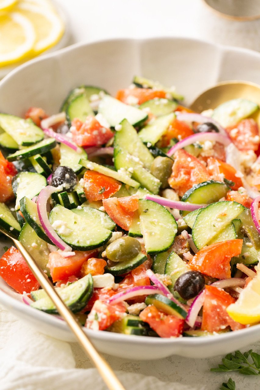 a closeup shot of greek cucumber salad with Mediterranean dressing in a white bowl with a gold serving spoon 