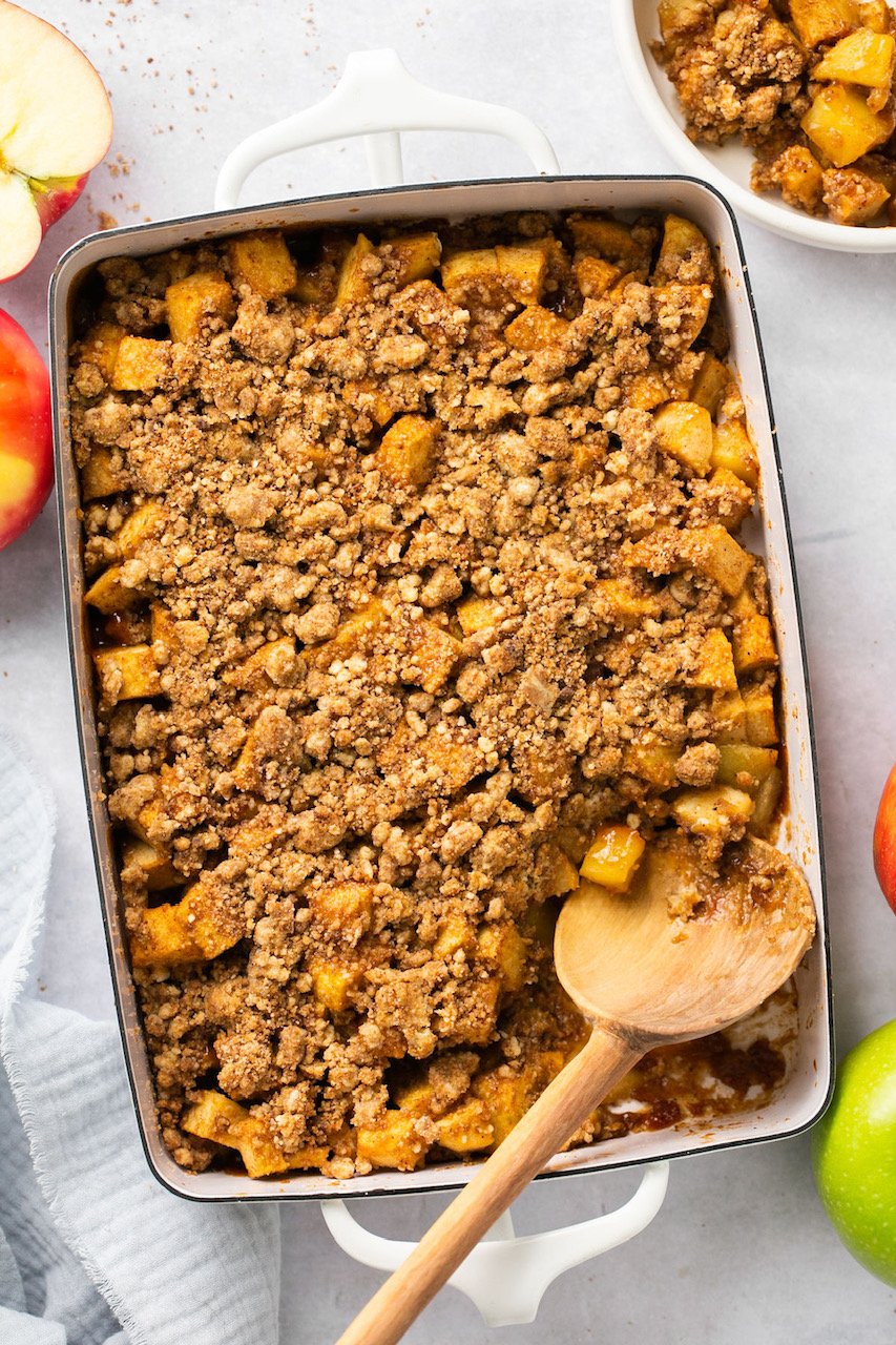 an overhead view of healthy apple crumble in a white casserole dish with a wooden spoon