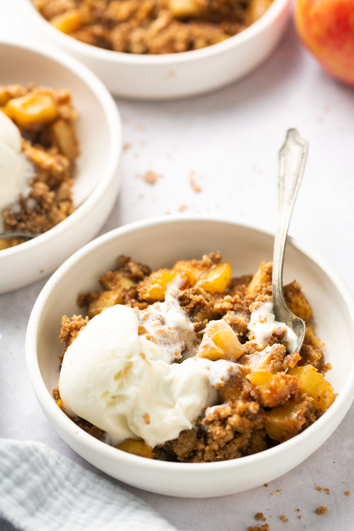 a slice of apple crumble in a white bowl topped with ice cream and a silver spoon 