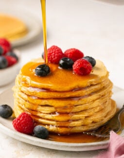 protein pancakes stacked on a plate with berries and maple syrup being poured on top