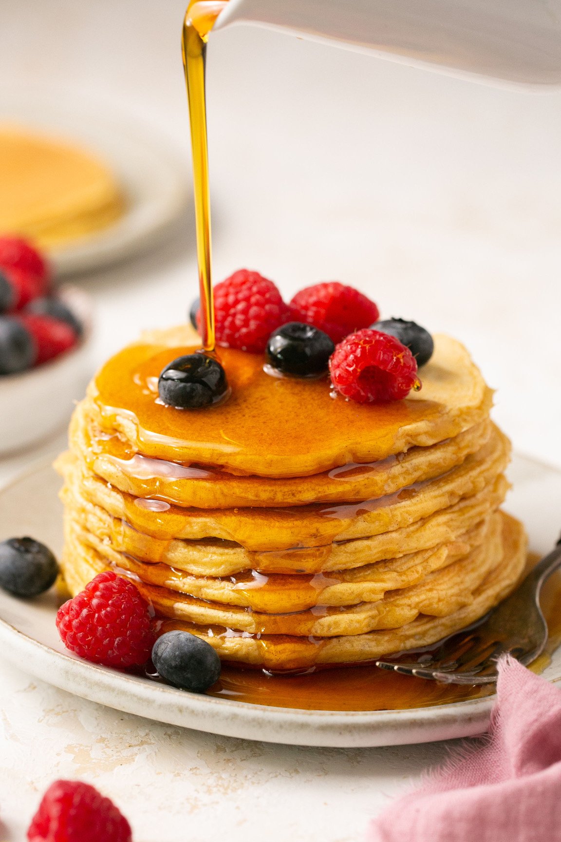 protein pancakes stacked on a plate with berries and maple syrup being poured on top 