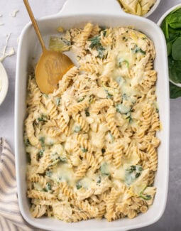 spinach artichoke chicken pasta bake in a casserole dish with a serving spoon taking a scoop out