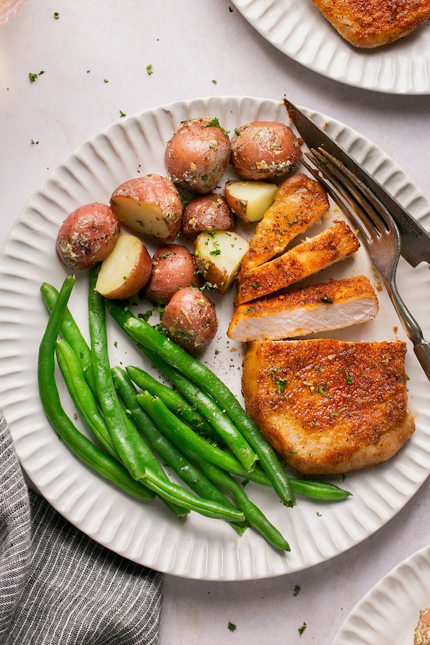 a white plate with roasted potatoes, green beans, and air fried pork chops with a fork and knife resting on the side