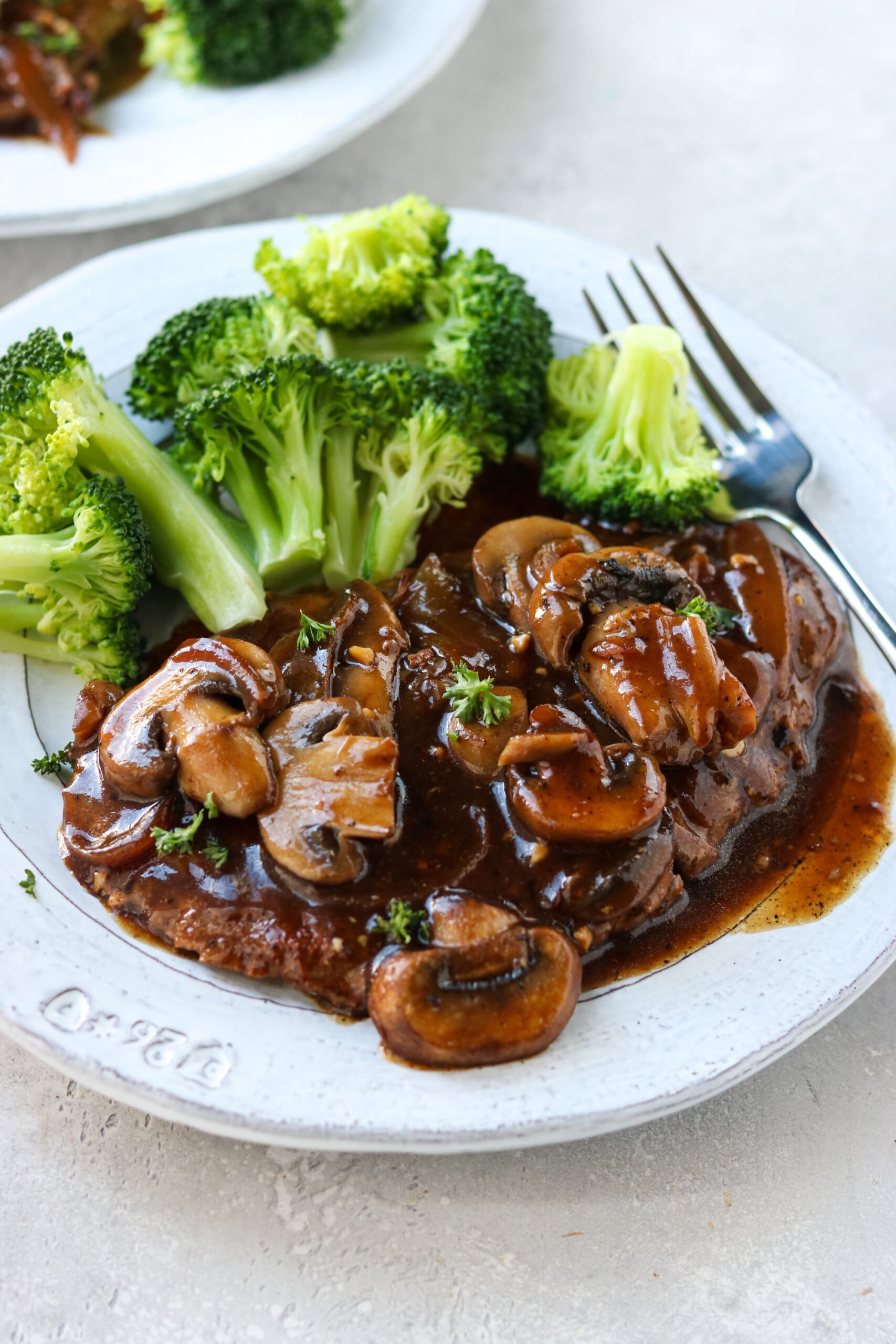 cube steak with gravy on a plate topped with mushrooms and next to a side of broccoli
