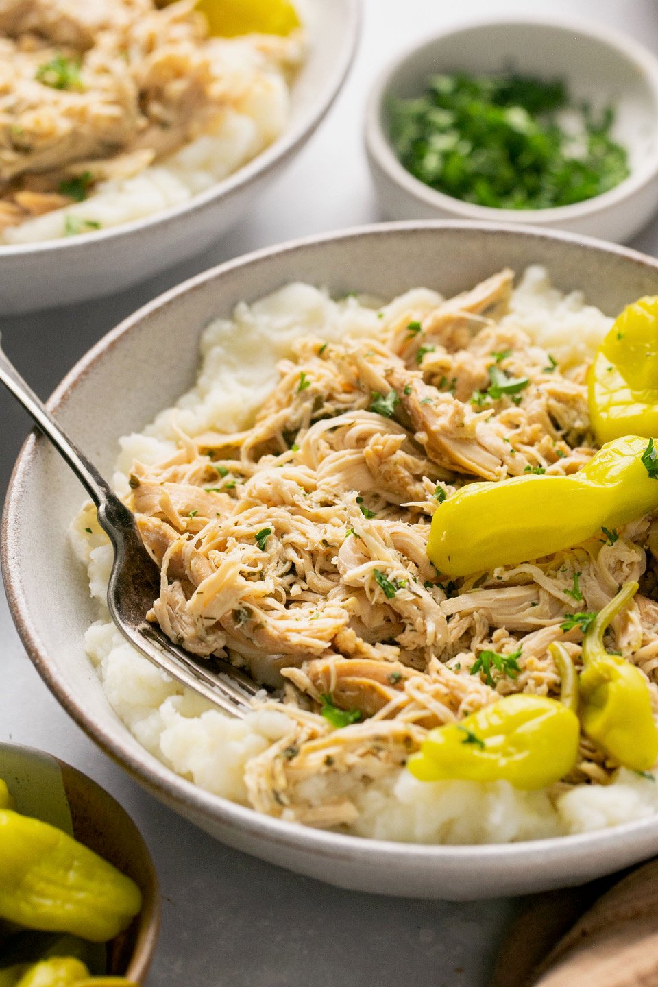 a white bowl filled with mashed potatoes, shredded chicken, and pepperoncini peppers