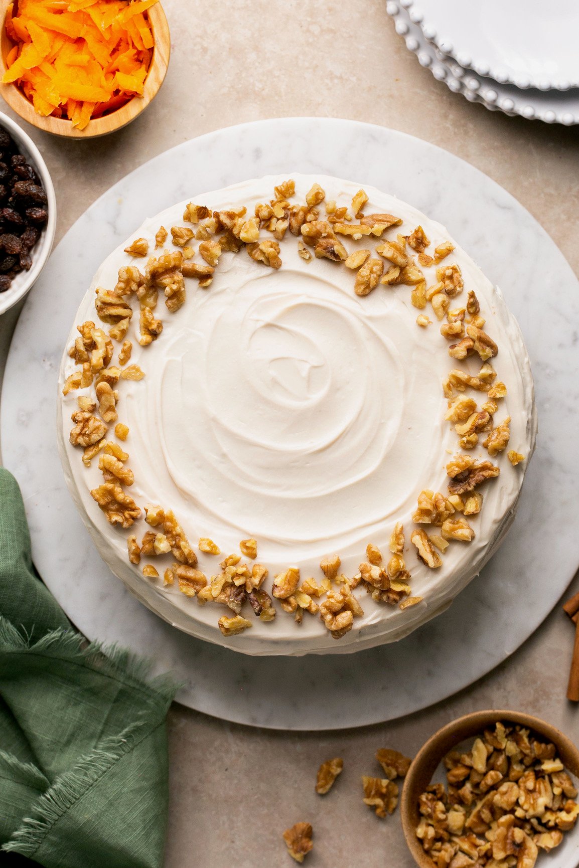 an overhead shot of carrot cake with walnuts surrounded by carrots, walnuts, a napkin, and plates.