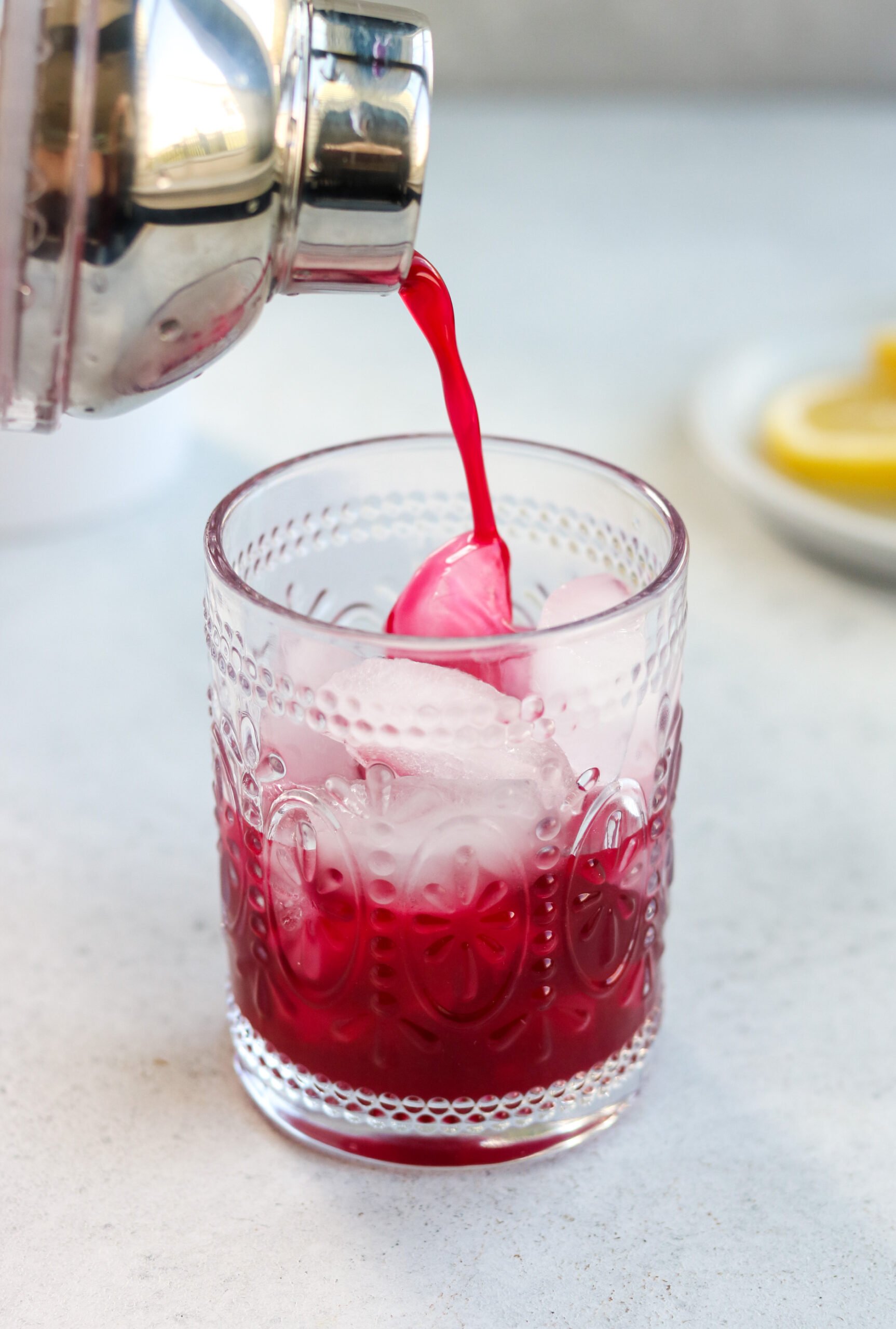 a cocktail shaker straining a lemon blueberry mocktail into a clear glass with ice cubes. 