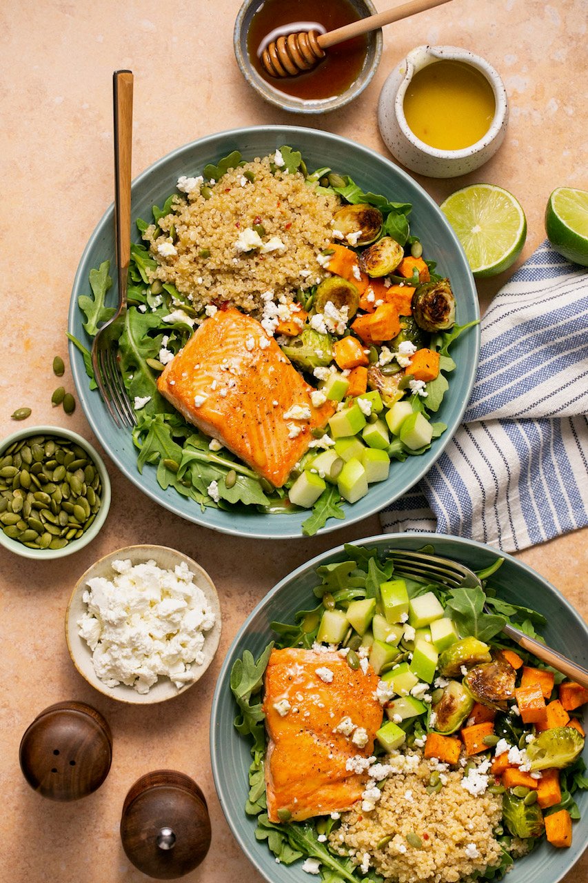 an overhead shot of two salmon quinoa bowls in blue dishes next to small bowls of additional ingredients. 