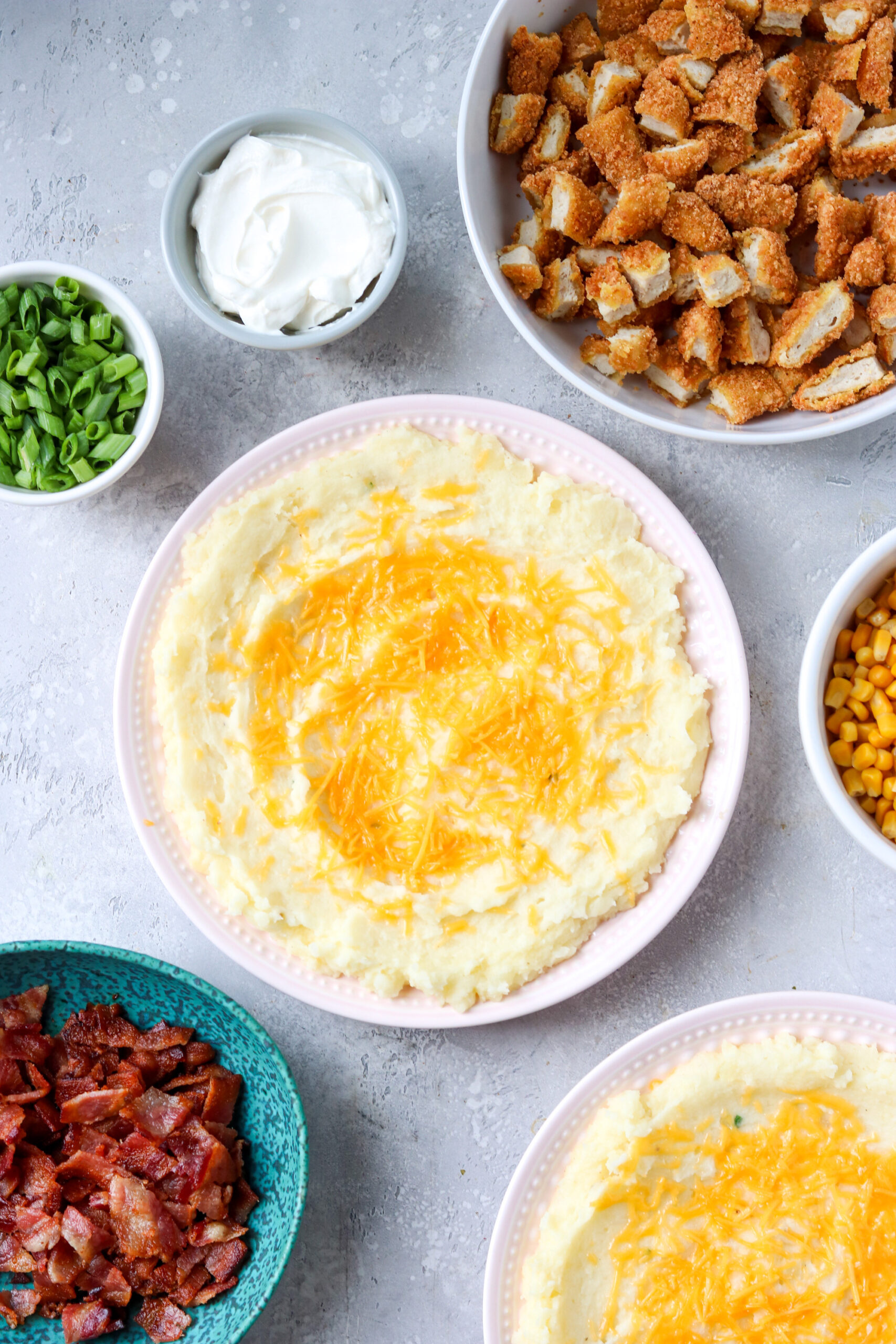 a bowl of mashed potatoes topped with cheese surrounded by bowls of chicken, sour cream, green onions, bacon, and corn