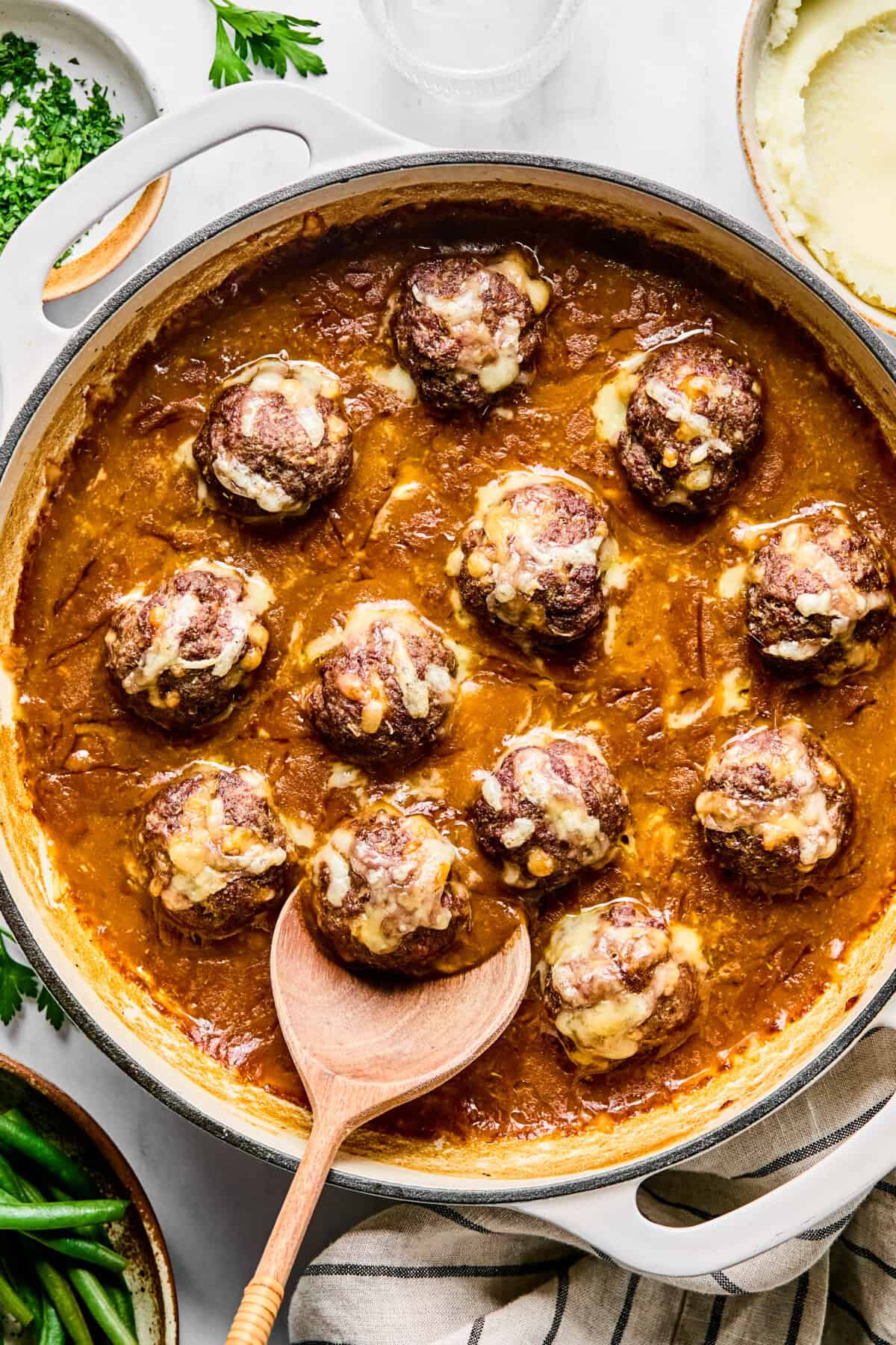 Baked French onion meatballs in a large oven-safe skillet; a wooden spoon scooping one out.