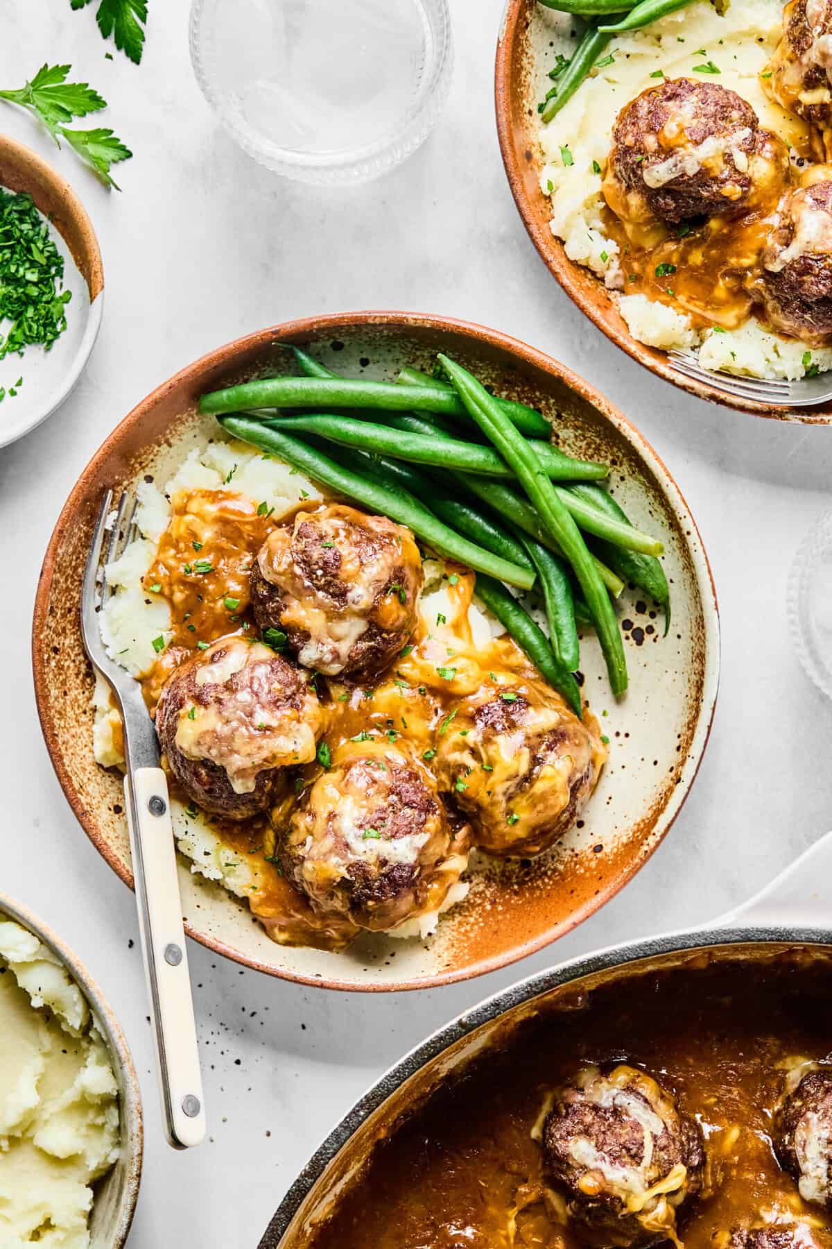 Plated French onion meatballs with onion gravy served over mashed potatoes and green beans on a light gray backdrop.