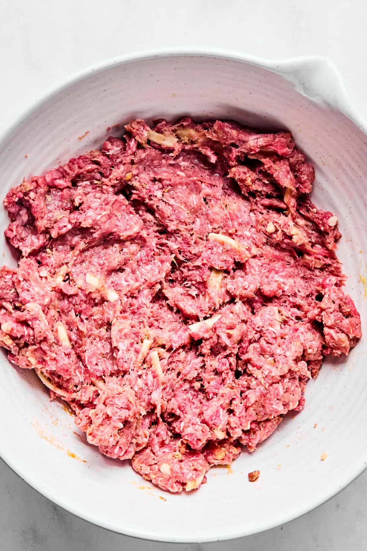 Ground beef, gluten-free flour, egg, seasonings, worcestershire sauce, and shredded cheese mixed in a white bowl.