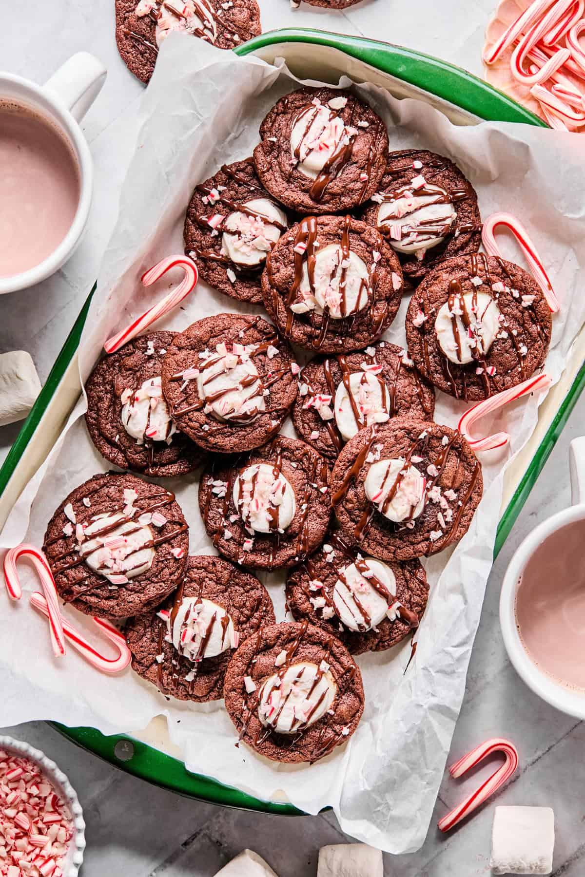 Gluten-free hot cocoa cookies arranged on a green parchment-lined tray, surrounded by mini candy canes with a mug of hot chocolate nearby.