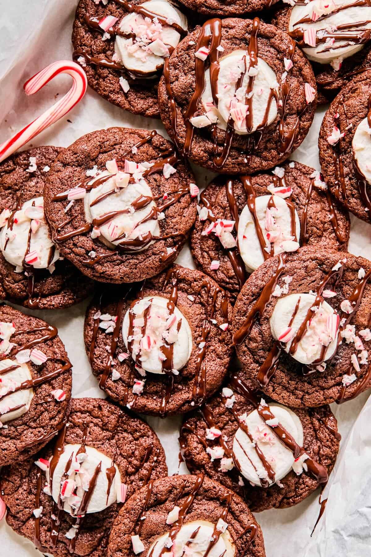 A batch of hot cocoa cookies (GF) stacked together on parchment paper topped with a chocolate drizzle and crushed candy canes.