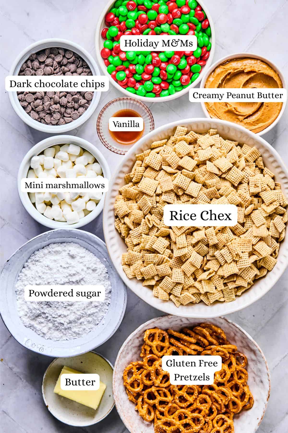Christmas puppy chow ingredients labeled and laid out in bowls including rice chex, powdered sugar, pretzels, peanut butter, vanilla, and chocolate chips.