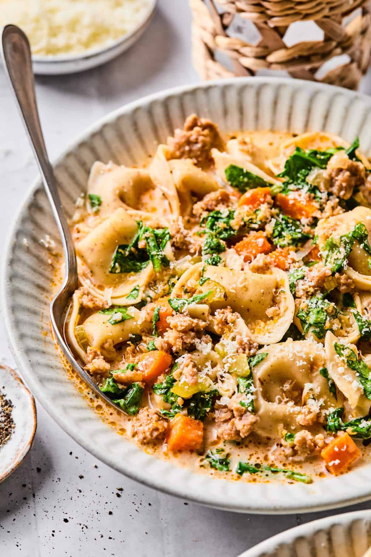 a close up of sausage tortellini soup with veggies, spinach and parmesan cheese on top. 