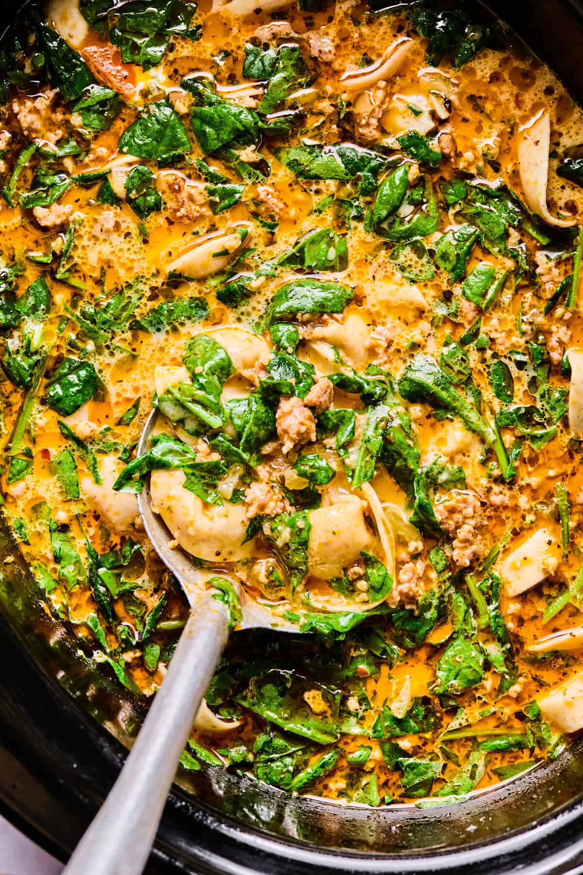 the finished italian sausage and tortellini soup in the crockpot with a large silver spoon 