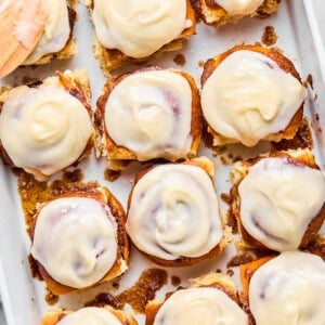 Hawaiian Roll Cinnamon Rolls in a baking dish with a spatula spreading a creamy glaze on one.