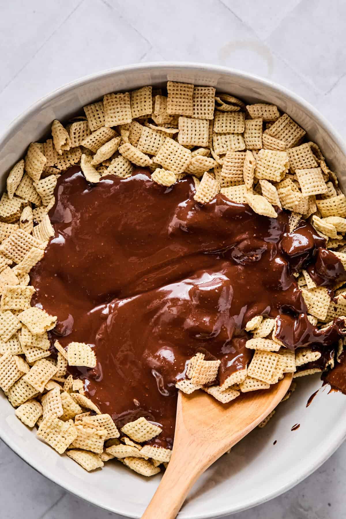 A large mixing bowl filled with Rice Chex cereal topped with melted chocolate and peanut butter, with a wooden spoon resting inside ready to mix.