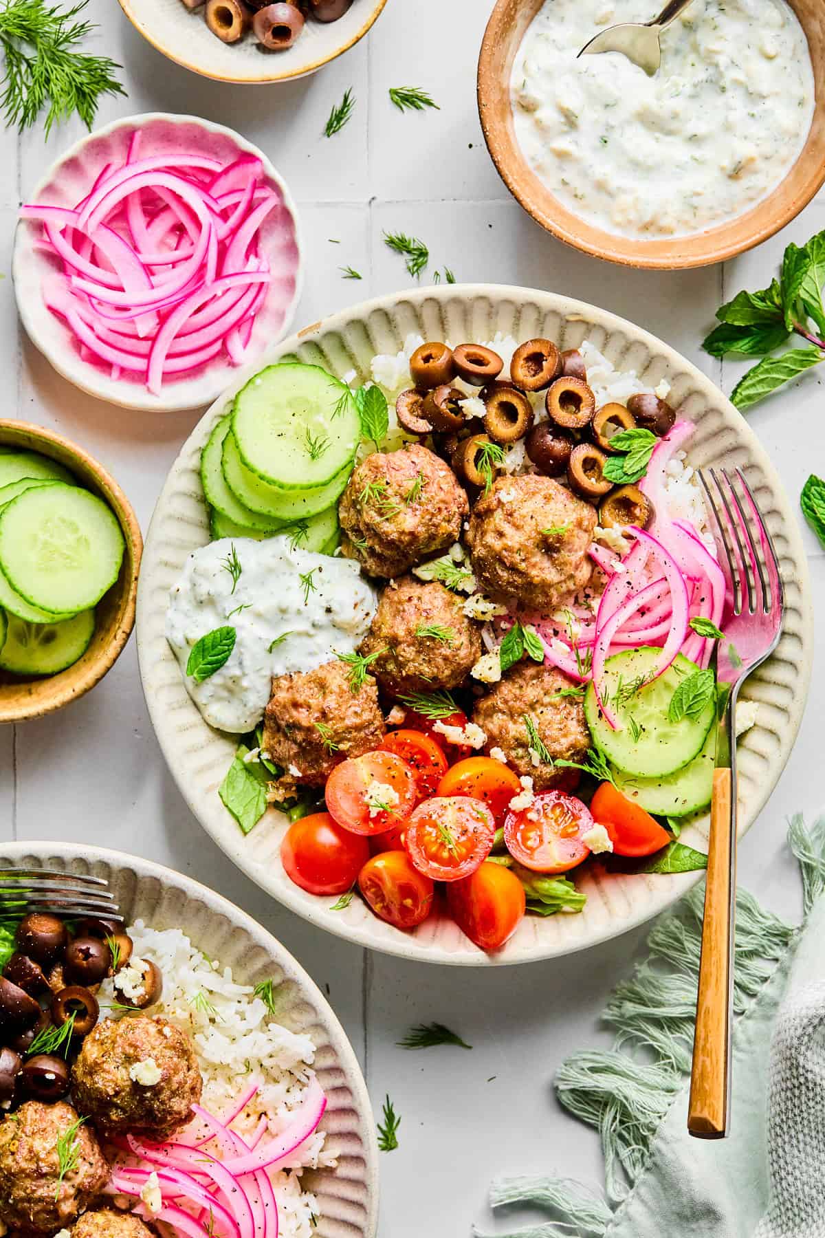 Greek lamb meatballs served over rice, surrounded by sliced cucumber, black olives, red onion, cherry tomatoes, fresh herbs, and crumbled feta cheese, with lemon dill yogurt sauce.