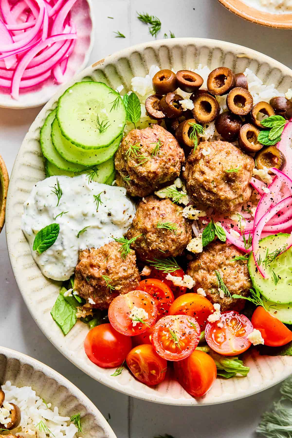 Close-up of Greek lamb meatballs served over rice, surrounded by sliced cucumber, black olives, red onion, cherry tomatoes, fresh herbs, and crumbled feta cheese, with lemon dill yogurt sauce on the side.