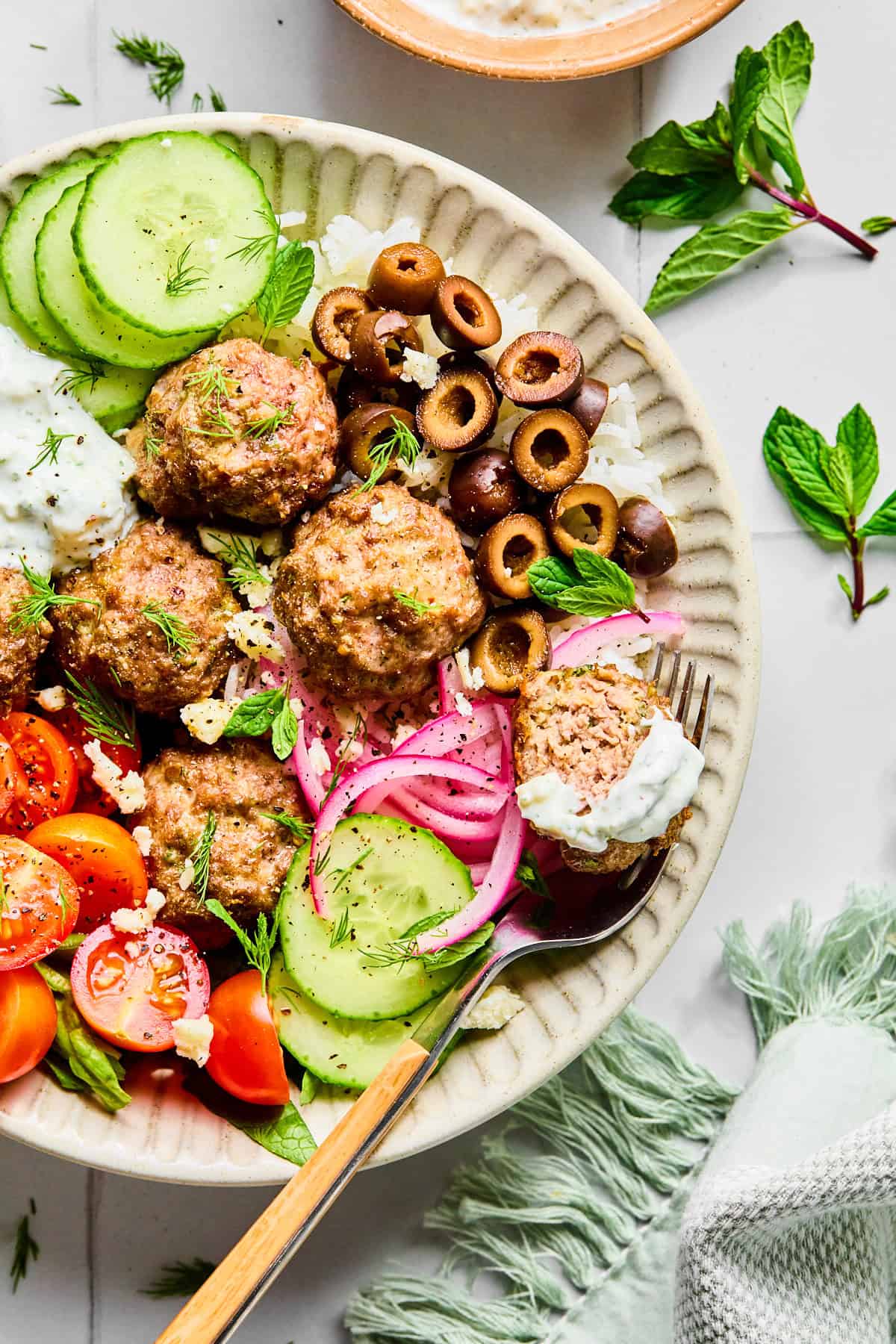 Greek lamb meatballs served over rice, surrounded by sliced cucumber, black olives, red onion, cherry tomatoes, fresh herbs, and crumbled feta cheese, with lemon dill yogurt sauce.