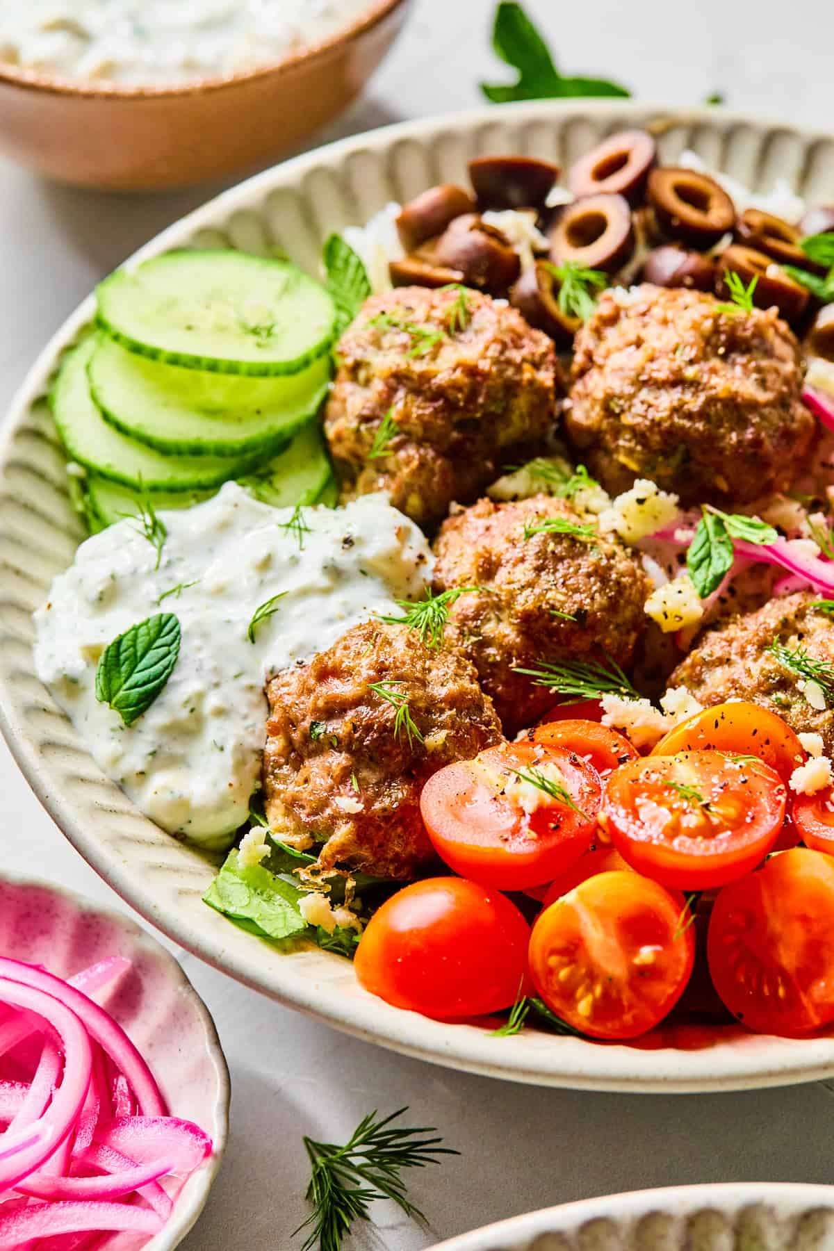 Side view of Greek lamb meatballs served over rice, surrounded by sliced cucumber, black olives, red onion, cherry tomatoes, fresh herbs, and crumbled feta cheese, with lemon dill yogurt sauce.
